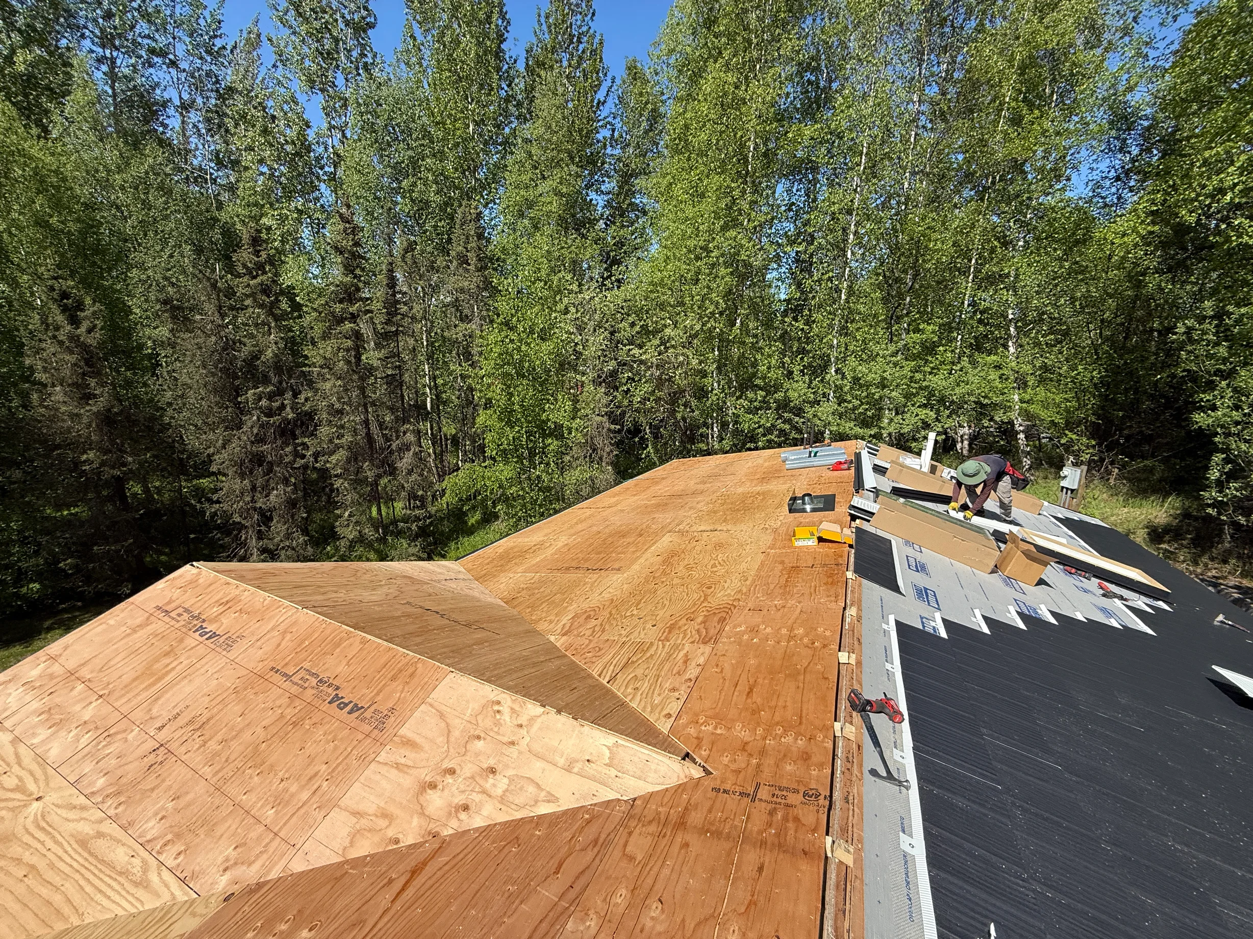 A modern-style house with a metal roof under construction, surrounded by lush greenery and a clear blue sky.