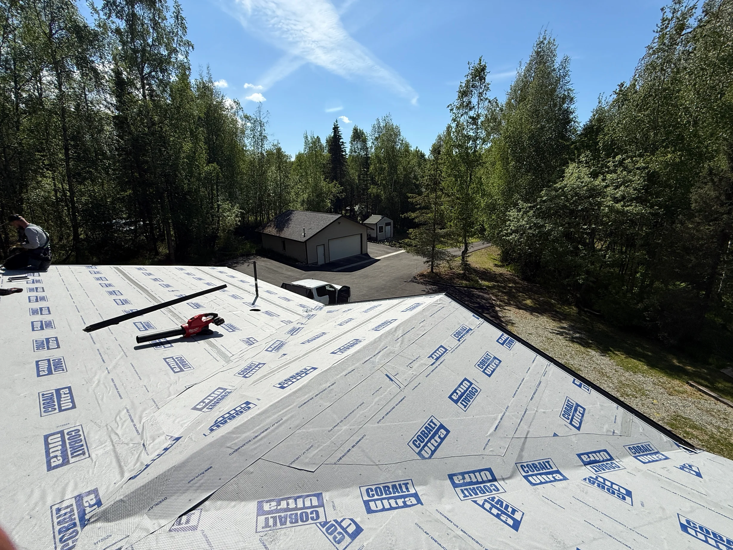 A metal roof is being installed on a house in a forested area on a sunny day.