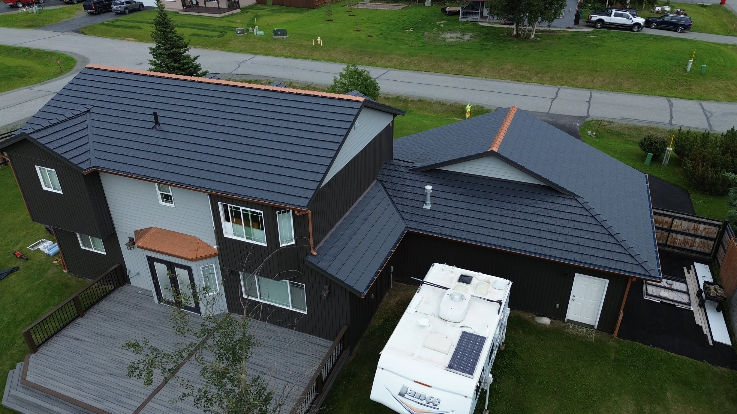 A modern single-family home with a metal roof and decking, located in an urban or suburban area with lush greenery and a blue sky.