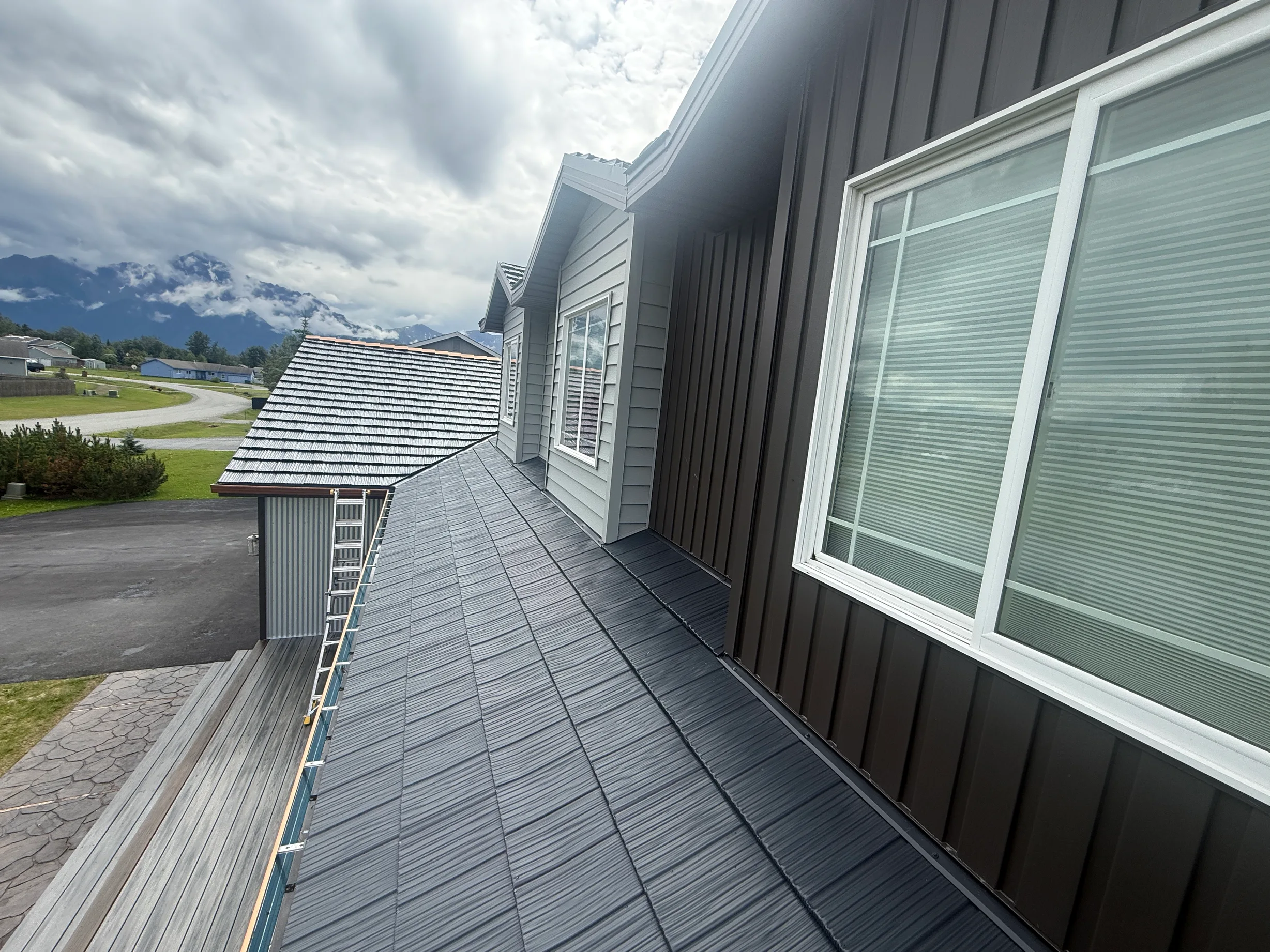 A modern home with a dark grey metal roof, set against a lush green mountain backdrop under an overcast sky.