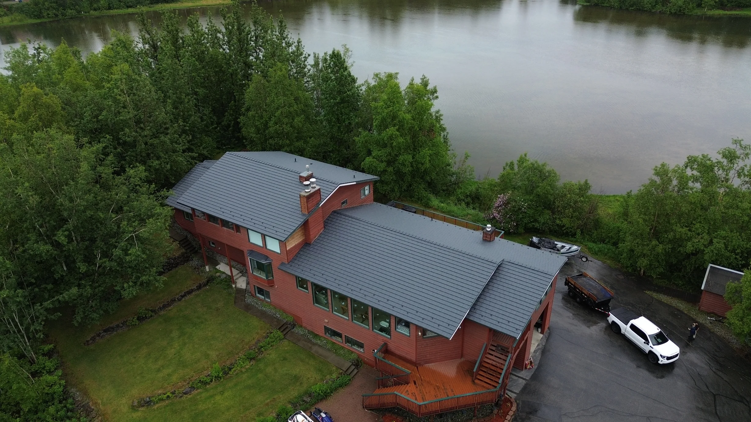 A modern single-family home with a metal roof and a chimney, overlooking a waterfront and forested area on a lush greenery day with overcast sky.
