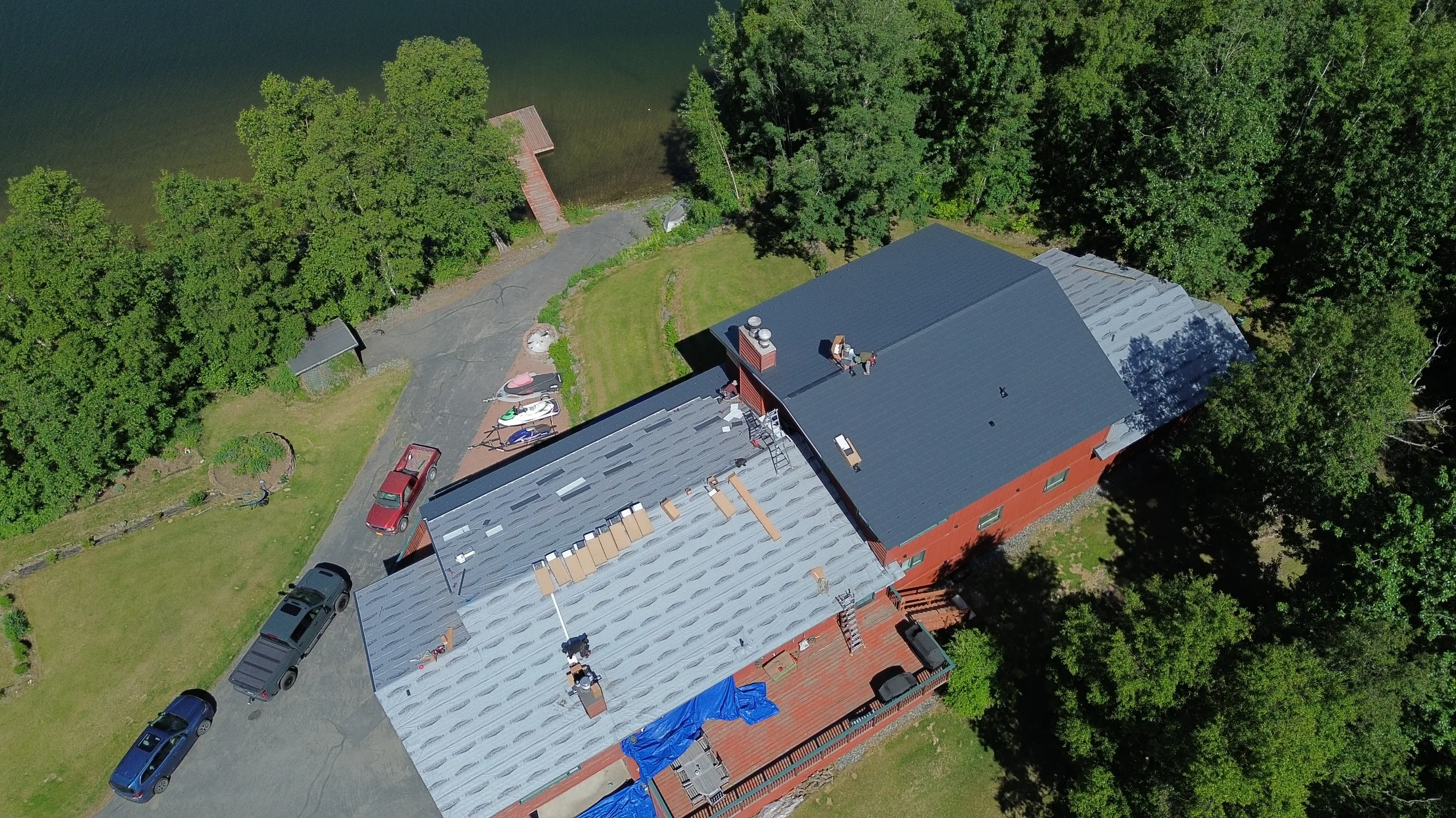Metal roof installation in progress on a large, red, brick house with grey shingles on a sunny day.