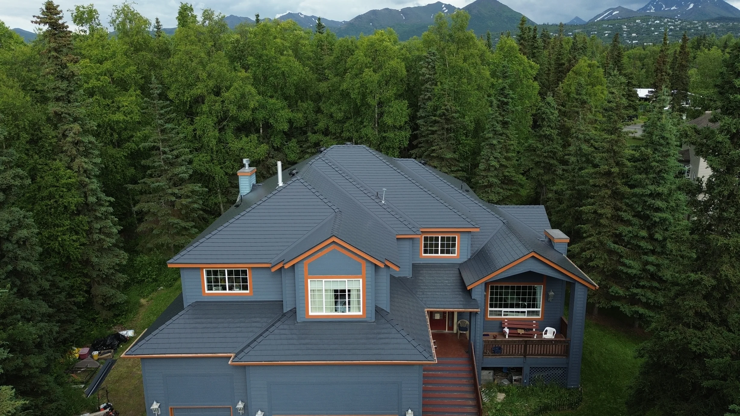A modern single-family home with a grey metal roof and brown trim, set against a lush forested backdrop with a mountain range in the distance.
