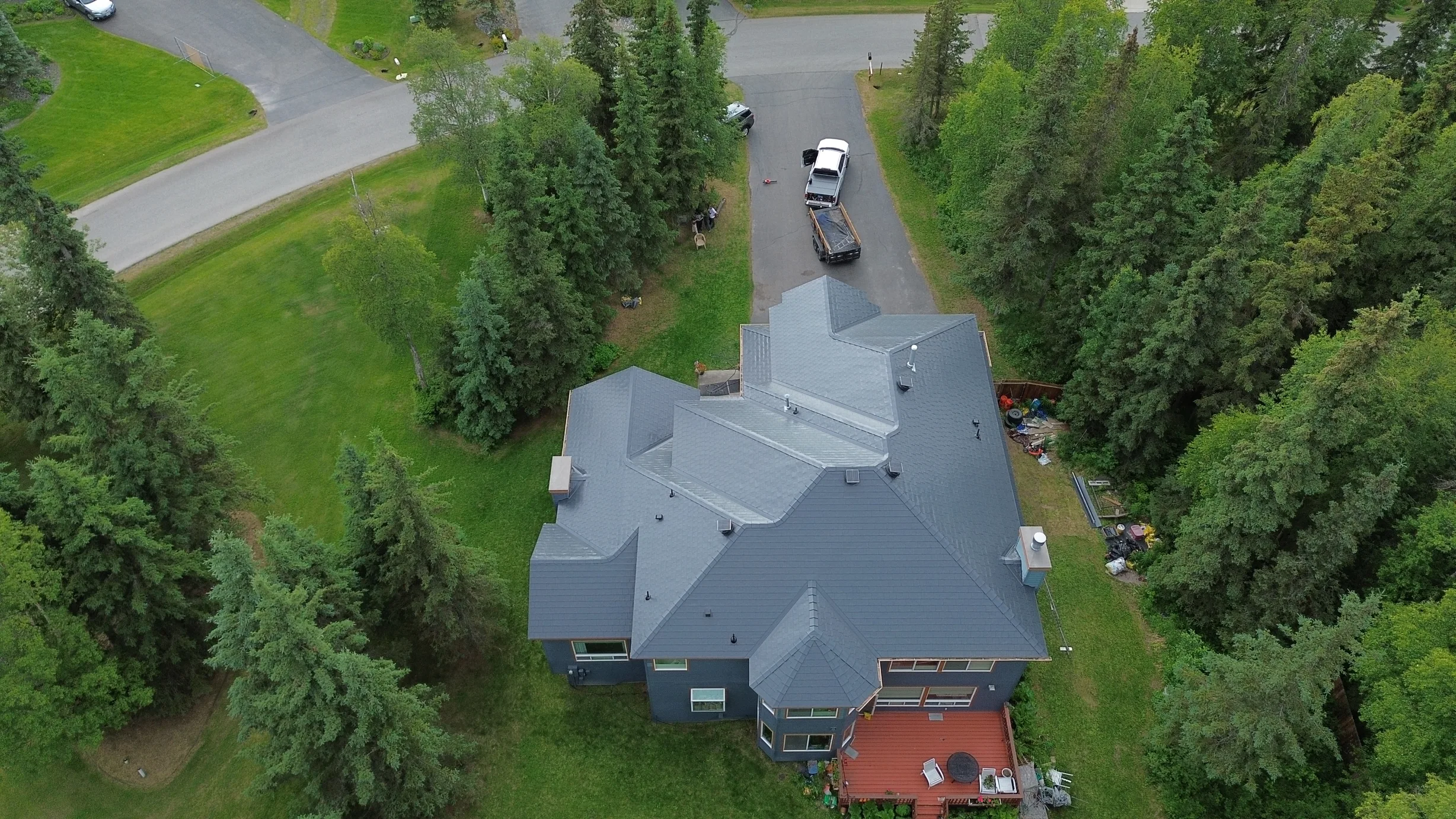 Aerial view of a modern single-family home with a grey metal roof, surrounded by lush greenery and trees.