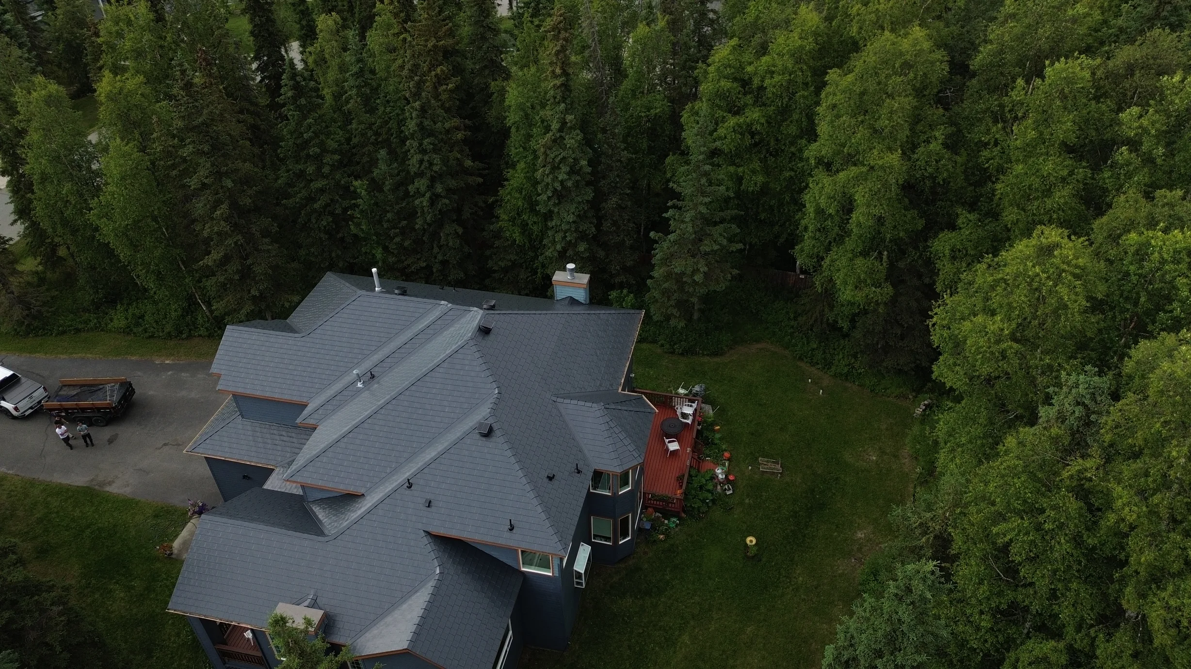 Aerial view of a modern single-family home with a grey metal roof, surrounded by lush greenery in a forested rural area.