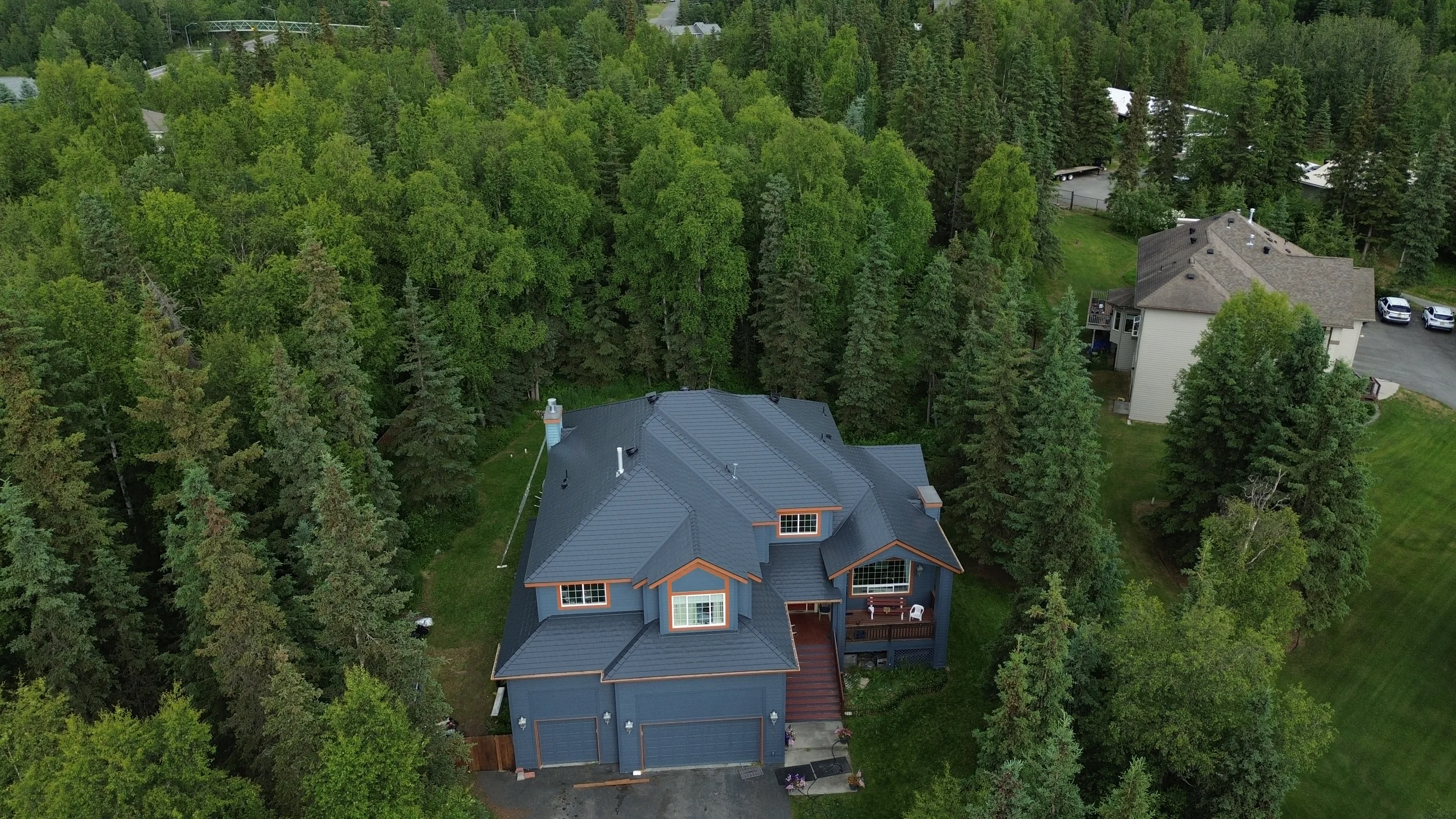 A large Colonial-style home with a grey metal roof and a chimney, set in a forested area surrounded by lush greenery and a clear blue sky.