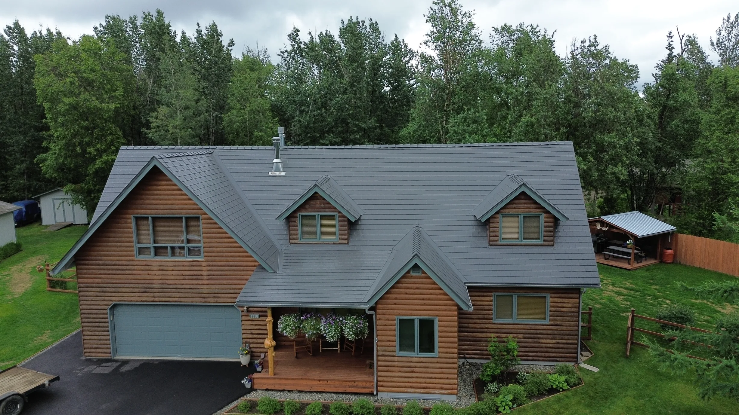A cottage with a grey metal roof and brown siding, surrounded by lush greenery and trees.