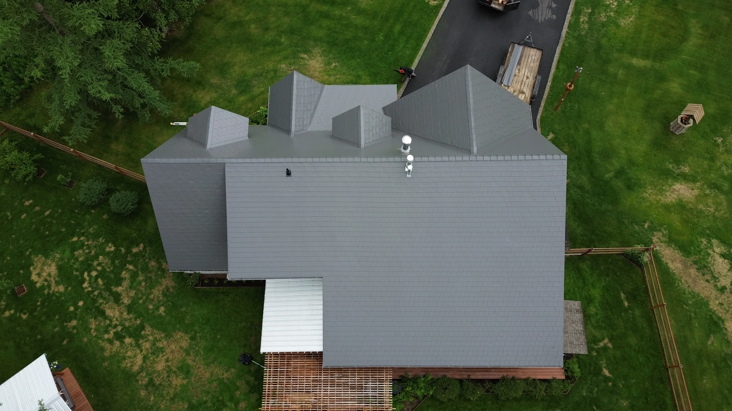 Aerial view of a modern, single-family home with a grey metal roof, snow guards and rails, and a roof louver, in a rural setting with lush greenery.