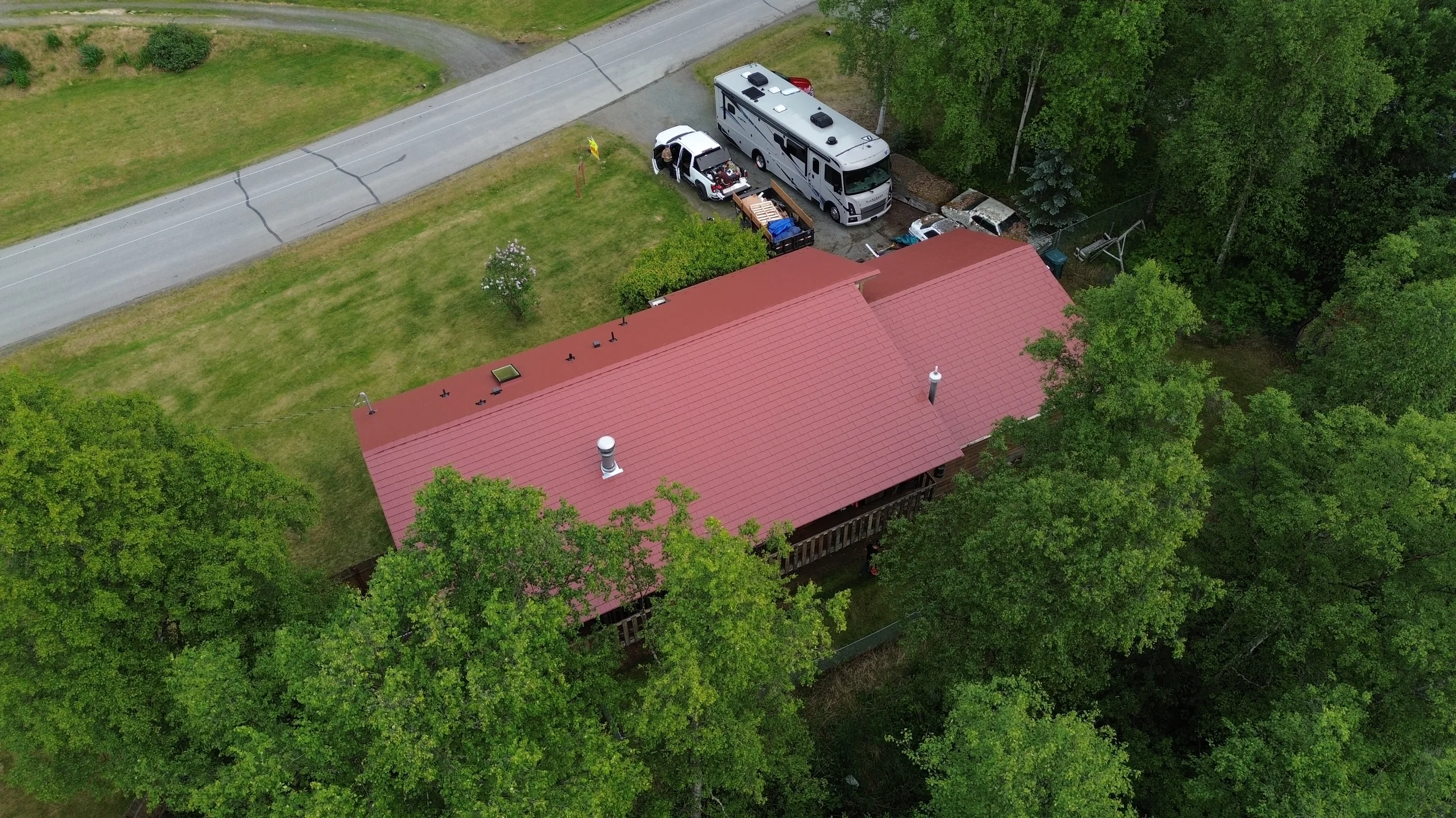 A ranch-style home with a red metal roof, surrounded by trees and a lush green lawn, with a few solar panels and a snow guard visible.