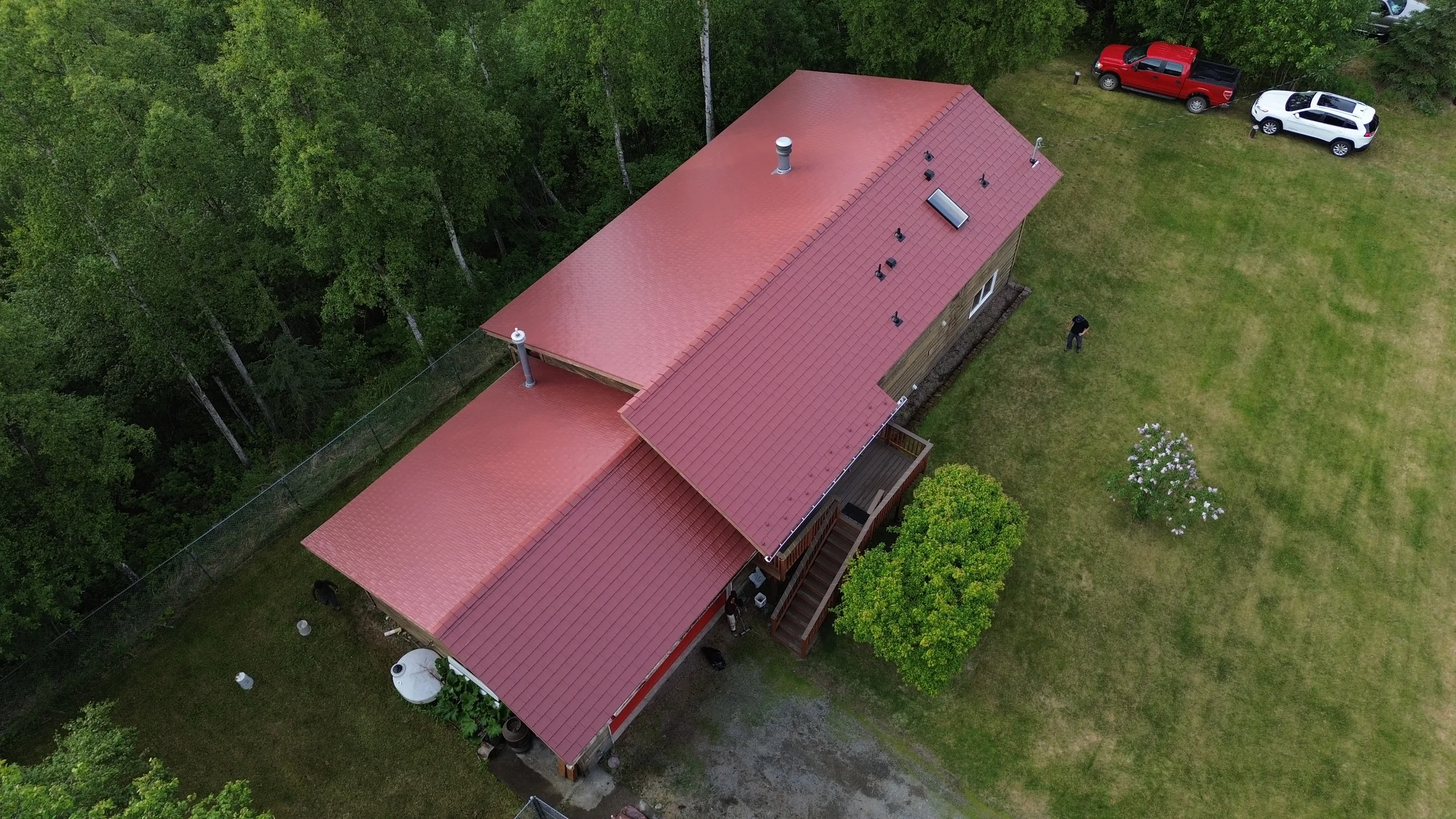 Aerial view of a ranch-style house with a red metal roof and solar panels in a wooded area