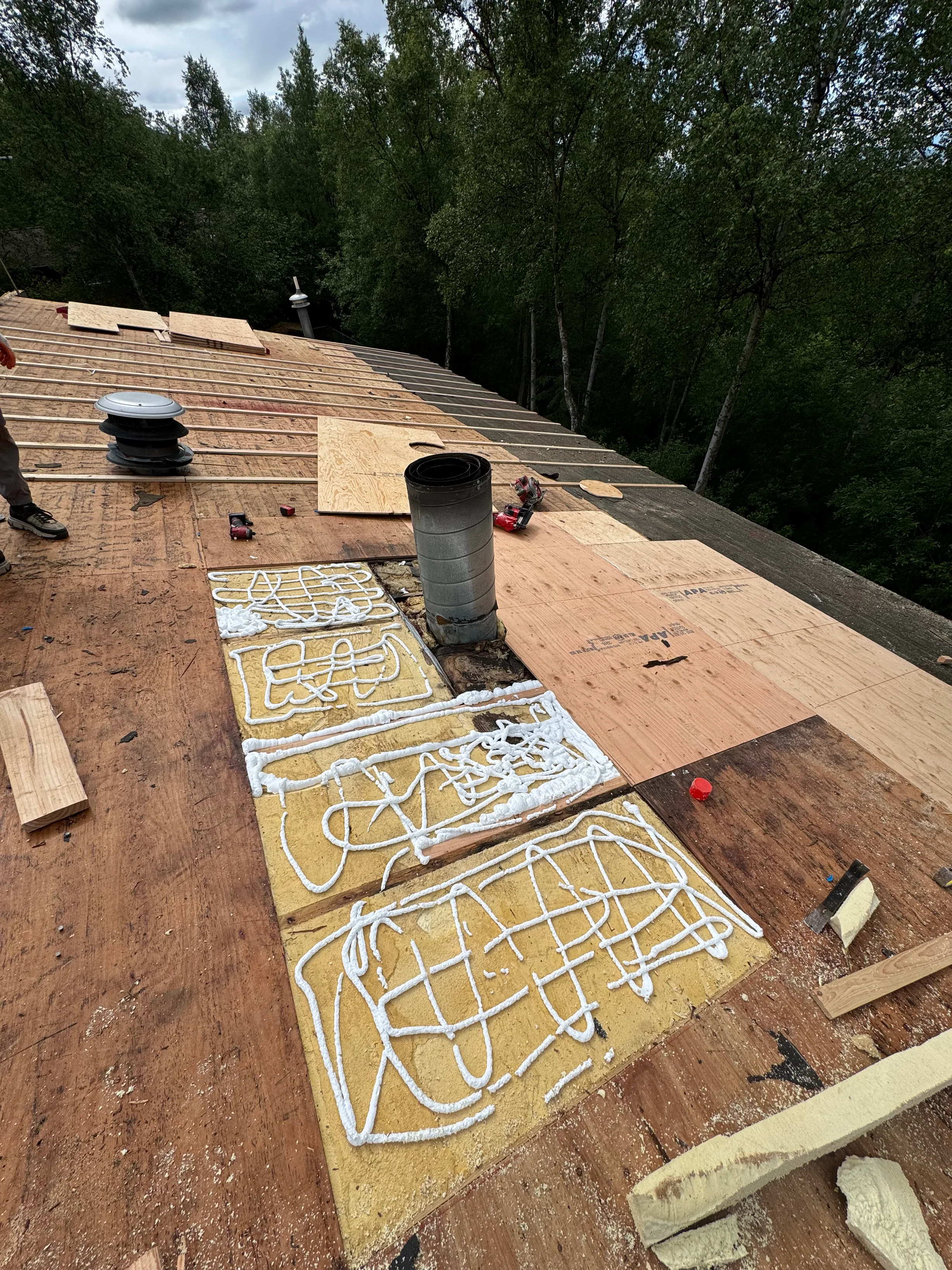 A colonial-style single-family home in a forested rural area with lush greenery and overcast skies, during installation of a metal roof.