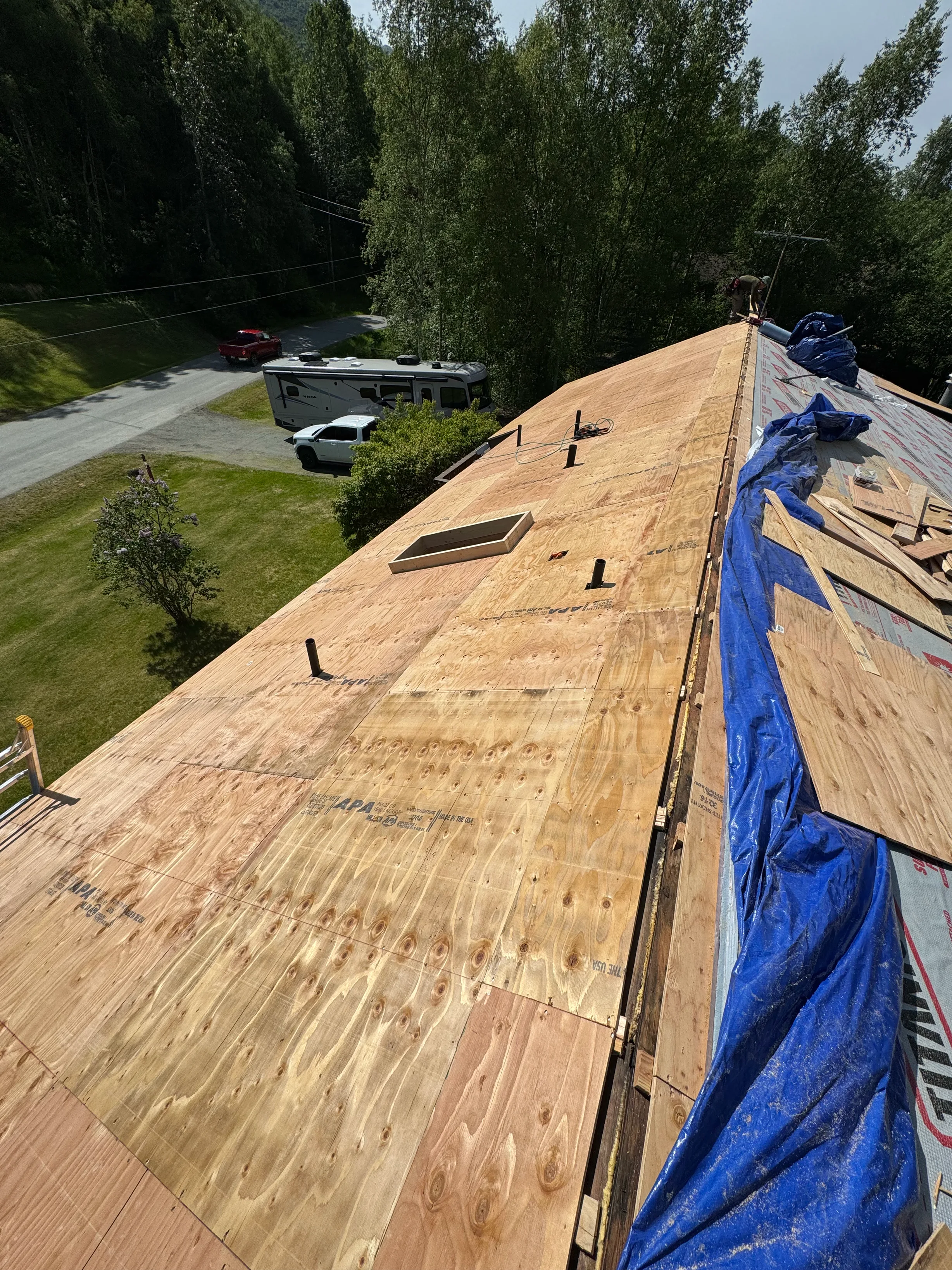 A partially installed metal roof on a ranch-style single-family home in a forested rural setting with a blue sky.