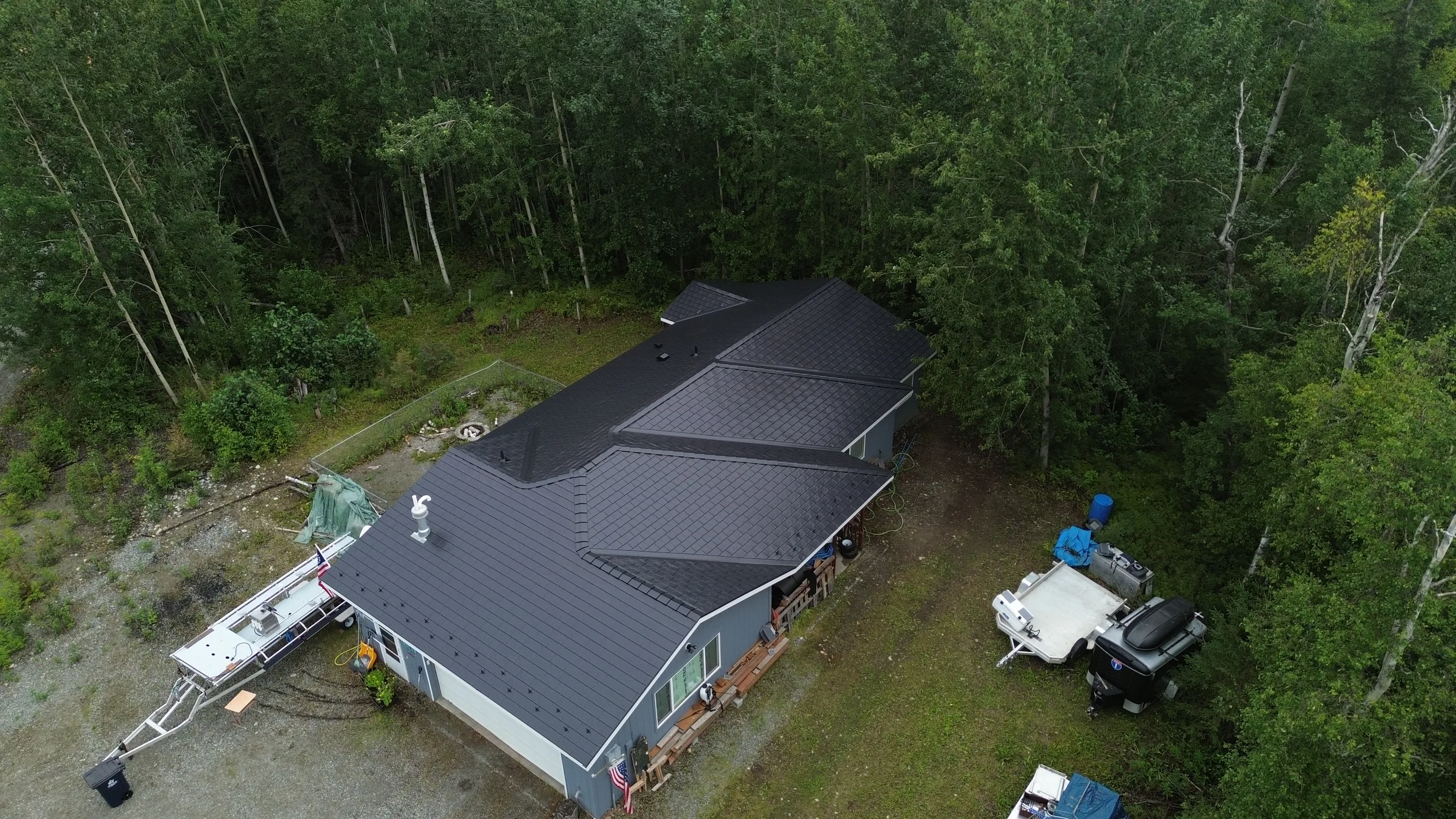 A metal roof with a dark grey colour on a single-family ranch-style home in a forested setting, with lush greenery and overcast conditions.