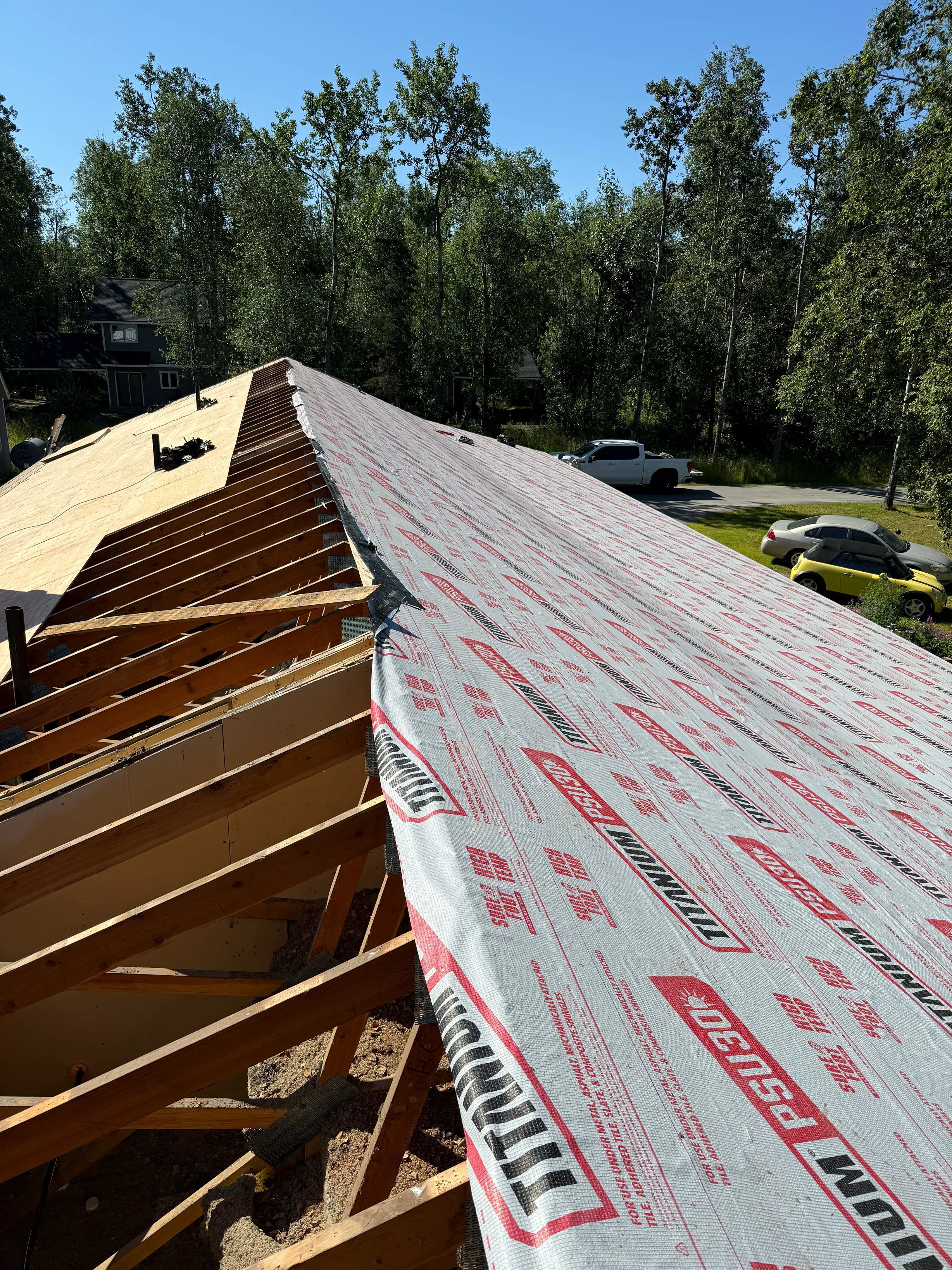 Metal roof installation in progress with ridge cap and snow guards visible, set against a lush greenery backdrop with a clear blue sky.