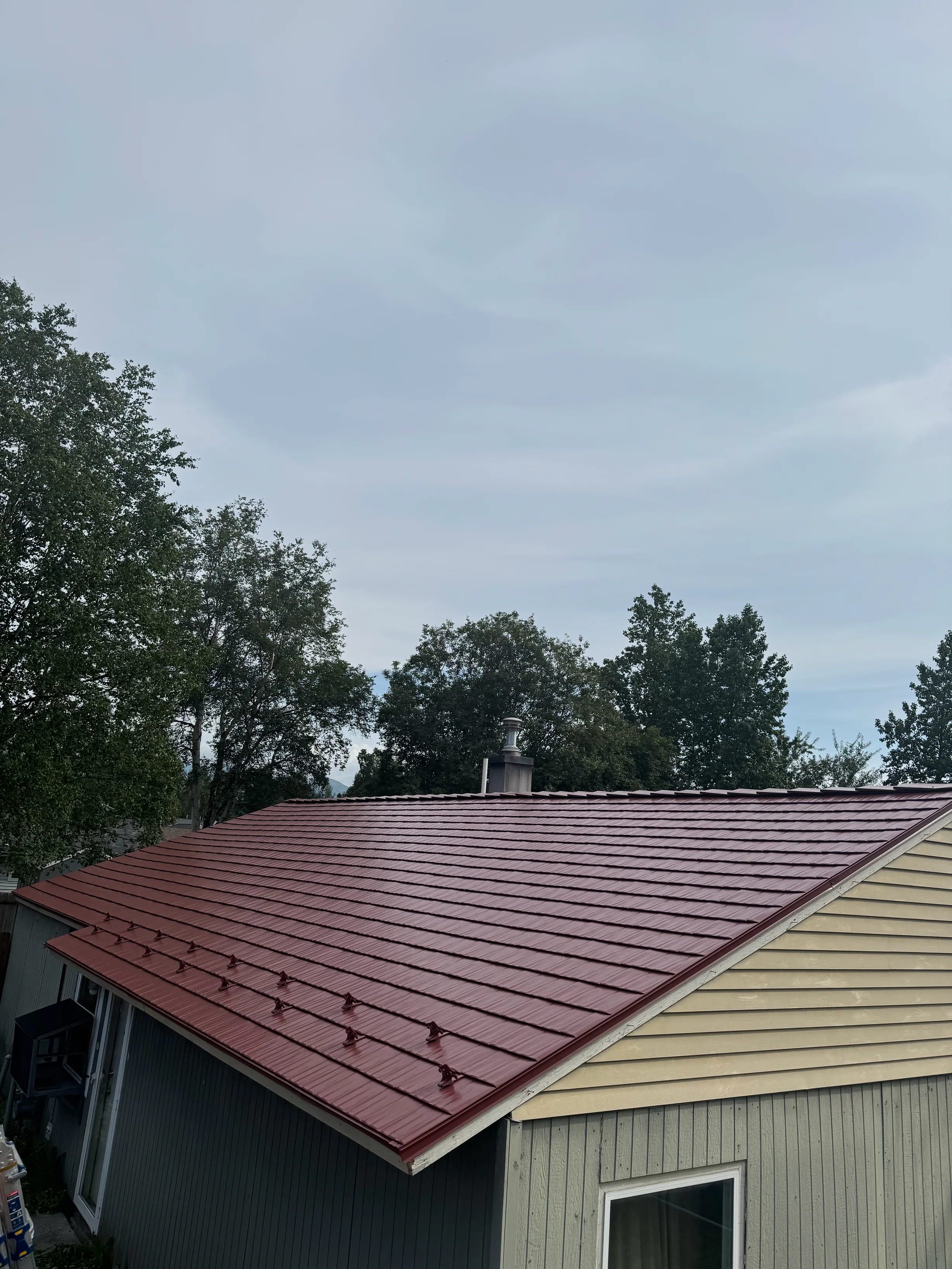 A modern single-family home with a red metal roof and lush greenery in the background.