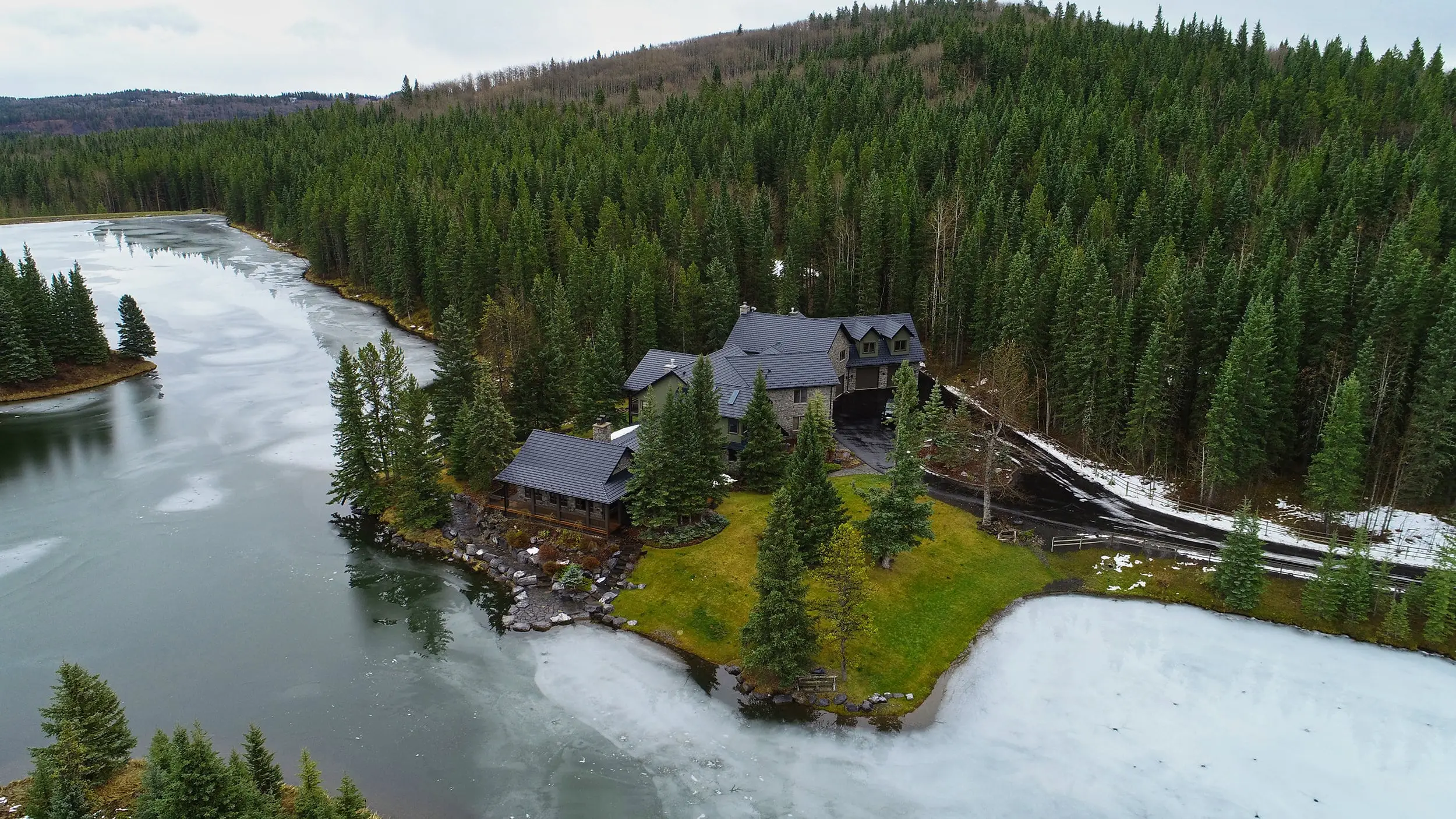 Aerial view of a chalet-style home on a lakeside peninsula in Bragg Creek AB featuring black Interlock aluminum shake metal roofing with complex multi-gable design.