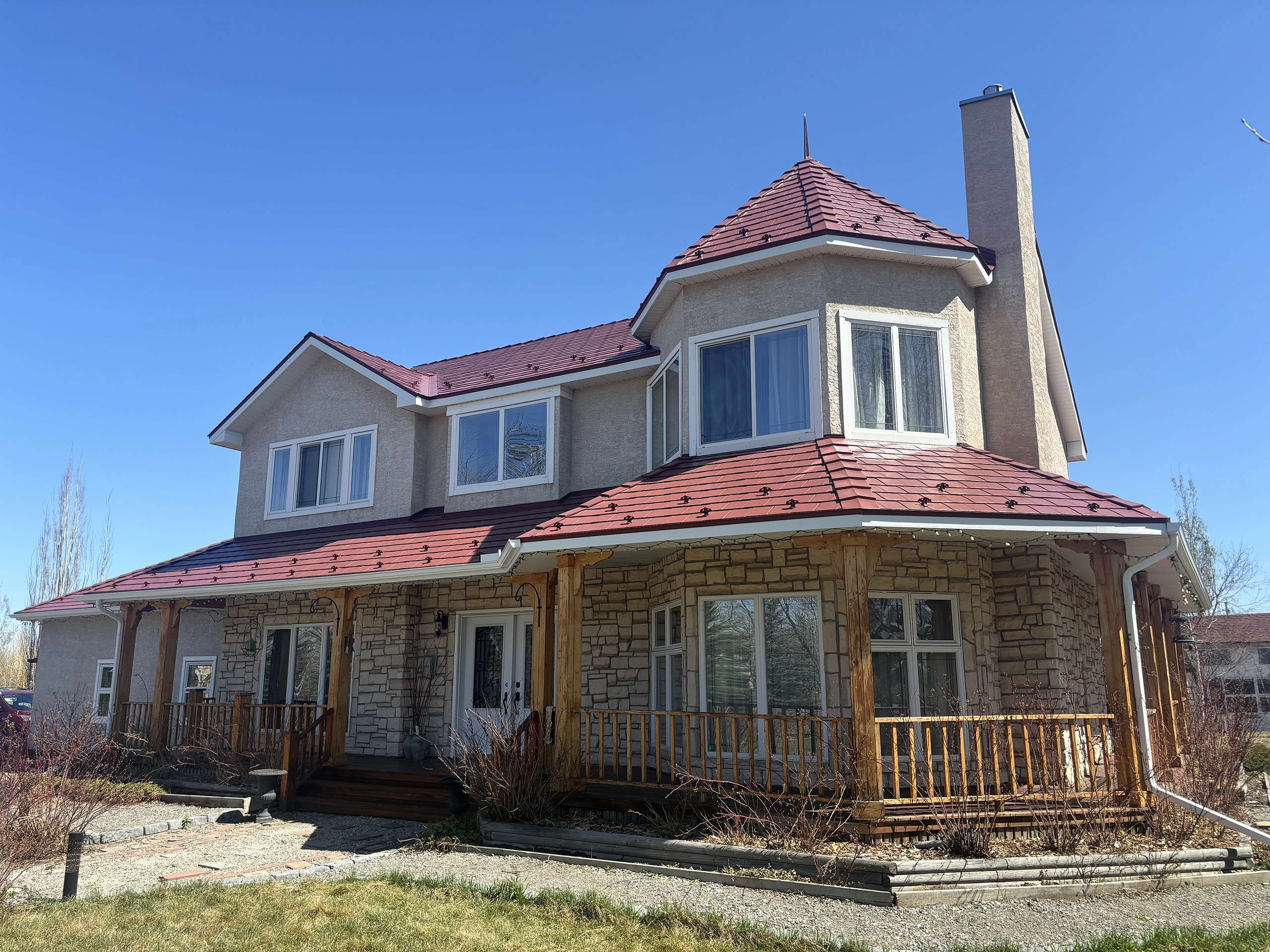 Two-storey stone-and-stucco home in Foothills County with a complex Tile Red Interlock aluminum slate metal roof featuring a turret and tall chimney.