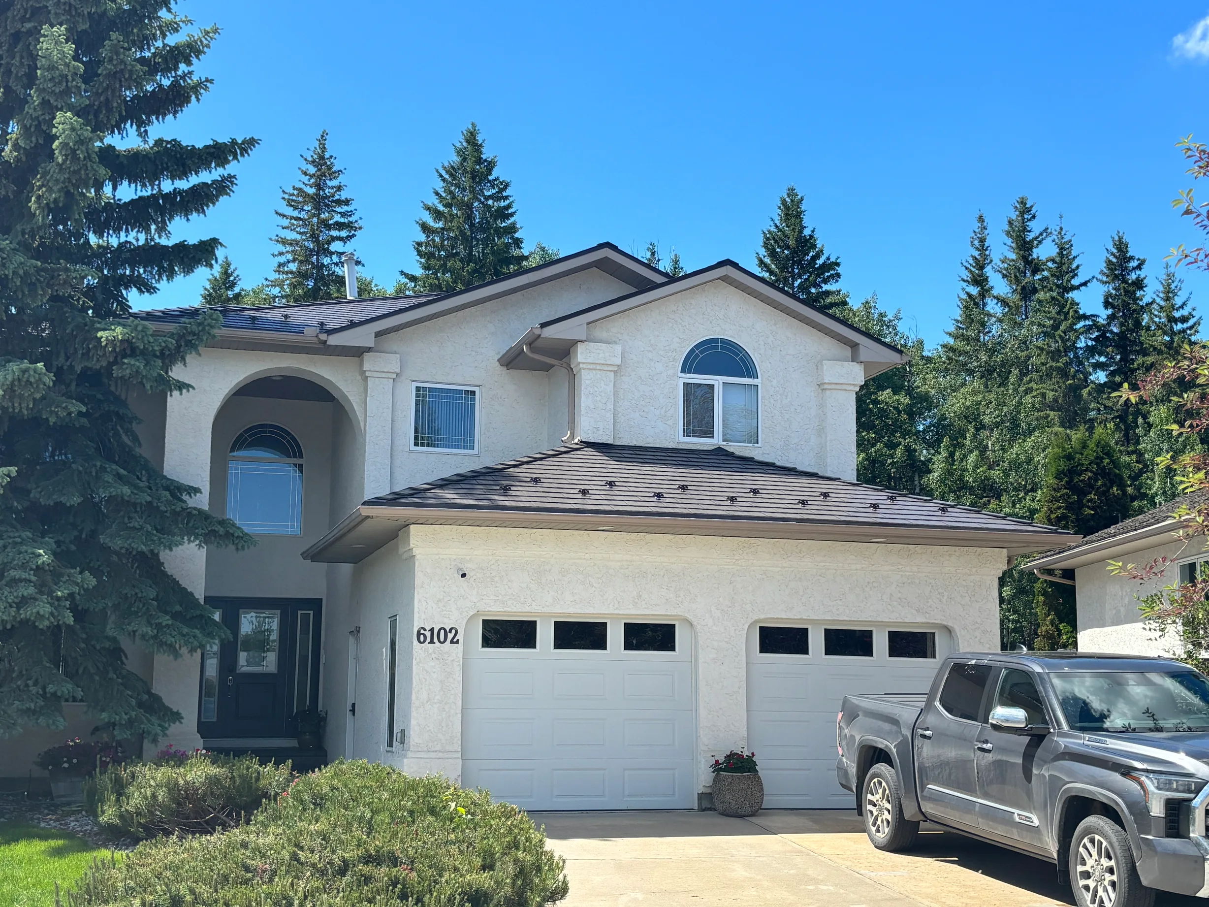 Tudor Brown Interlock Cedar Shingle aluminum roof on a white two-storey home with dormers among tall pines, Grande Prairie, Alberta