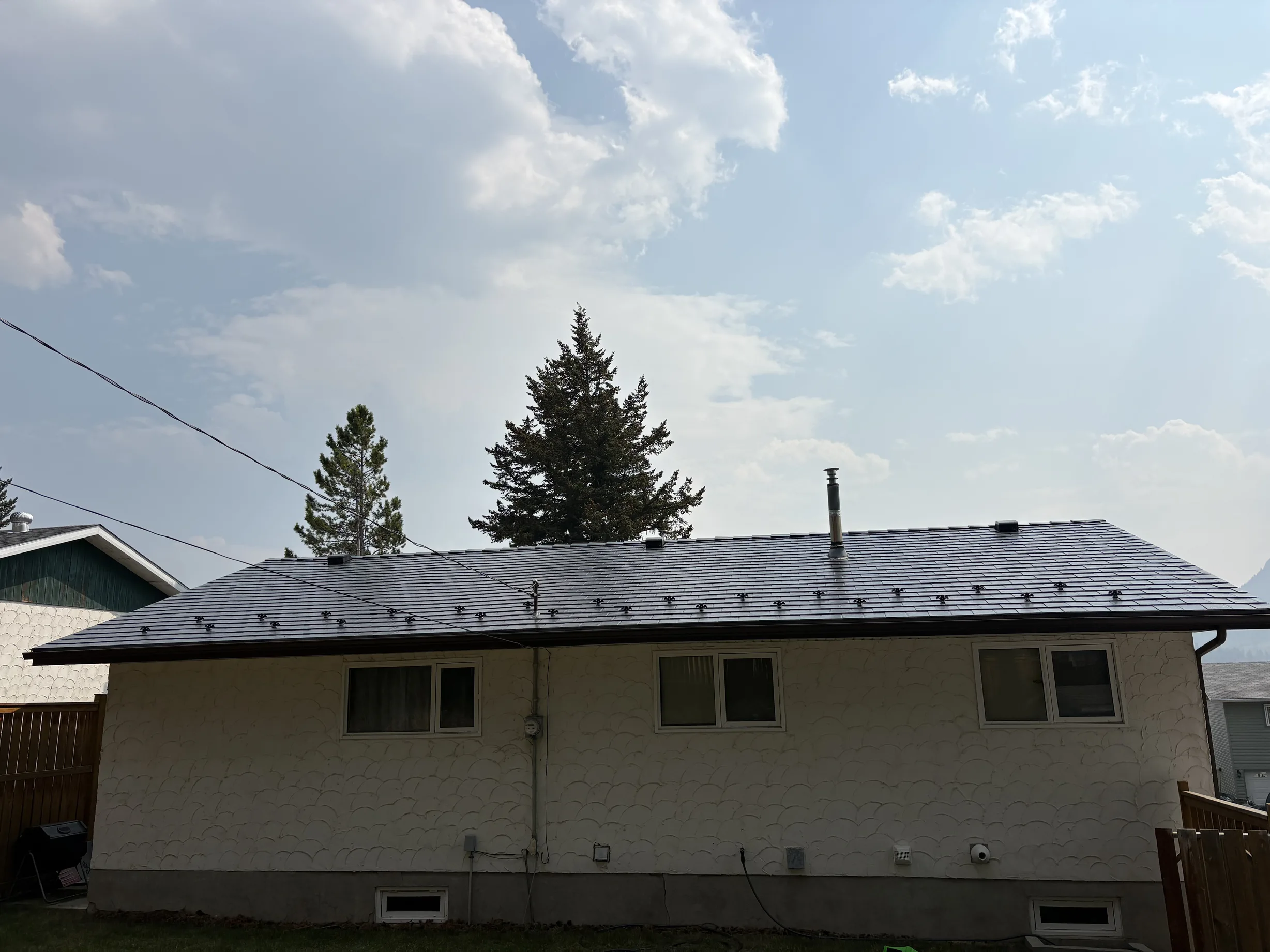 Deep Charcoal Interlock Slate aluminum roof on a white ranch-style home with a metal roof among tall trees, Coleman, Alberta