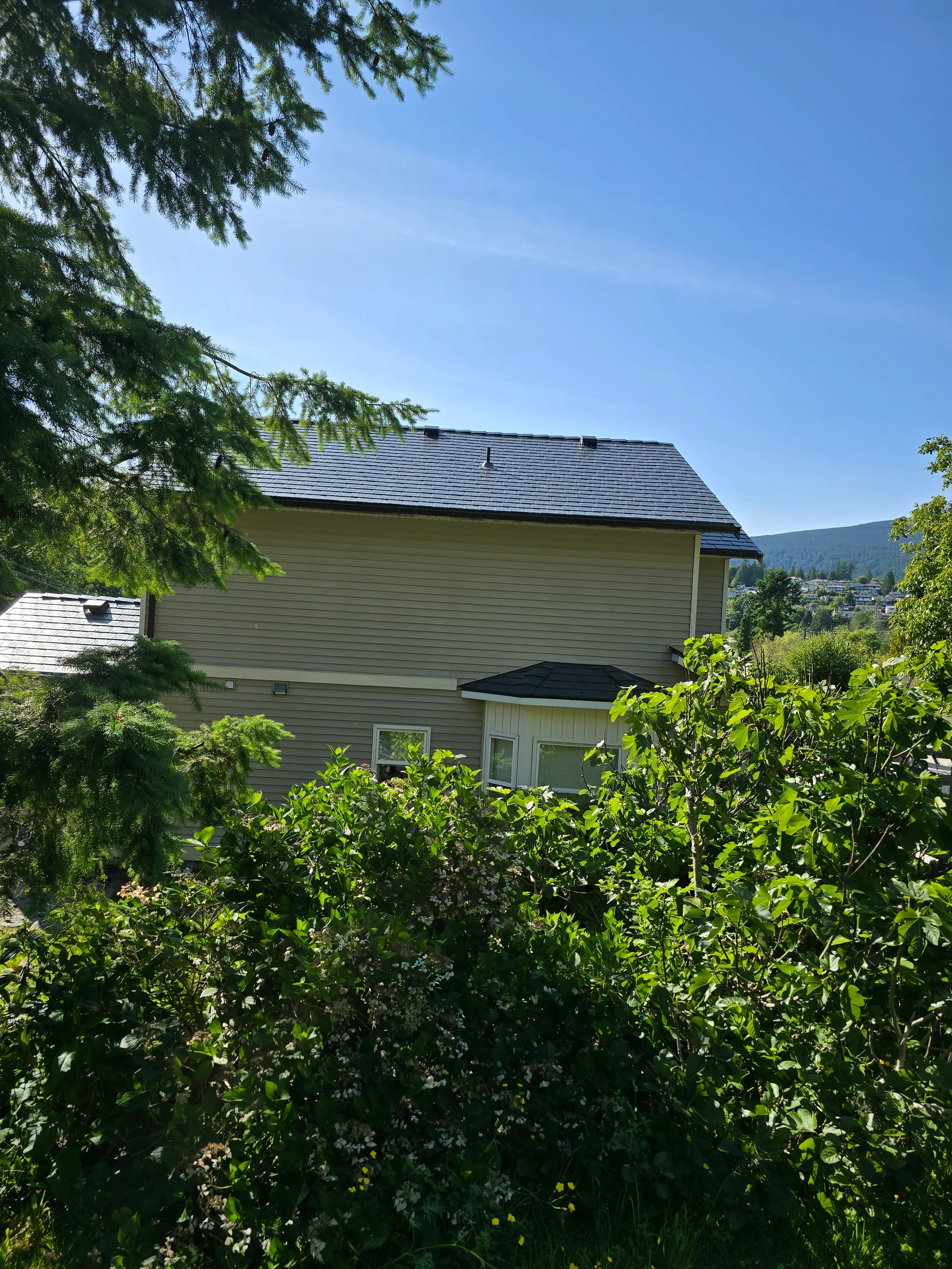 Deep Charcoal Interlock Slate aluminum roof on a tan two-storey home with dormers among tall pines, Gibsons, British Columbia