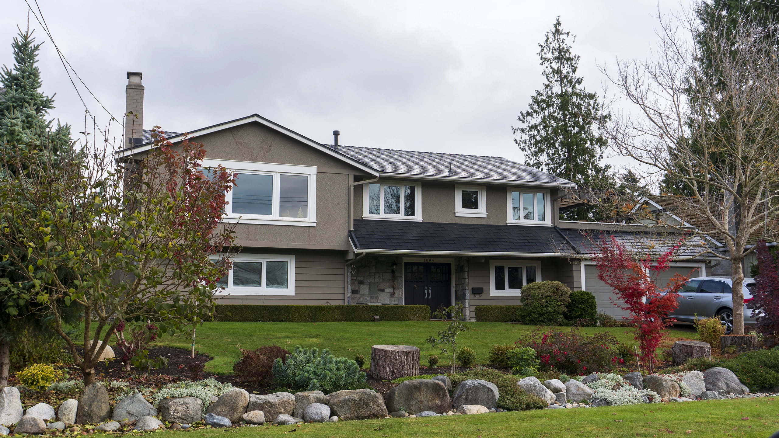 Two-storey suburban home in Tsawwassen BC with black Interlock aluminum shingle metal roof, stucco exterior, chimney, and landscaped front yard.