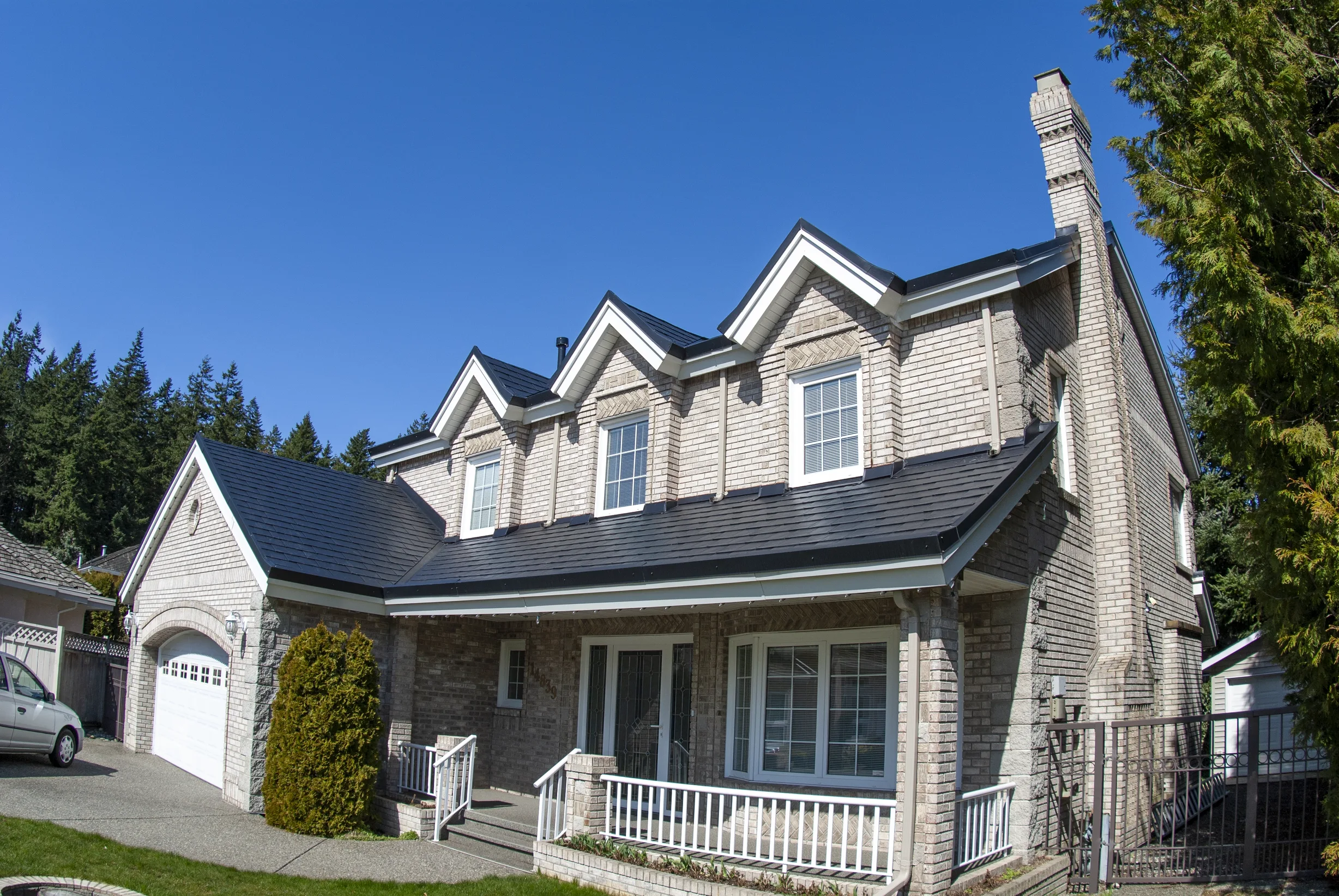 Black Interlock Slate aluminum roof on a beige two-storey colonial home with dormers among tall, Surrey, British Columbia
