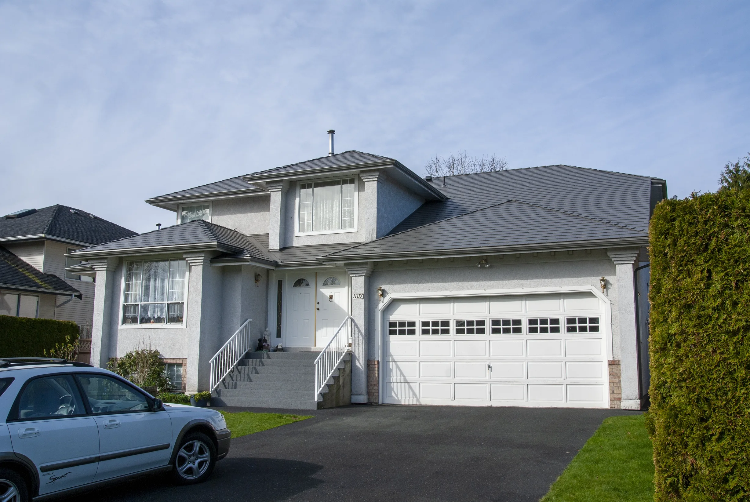 Musket Grey Interlock Slate aluminum roof on a white two-storey home with dormers among tall pines, Surrey, British Columbia