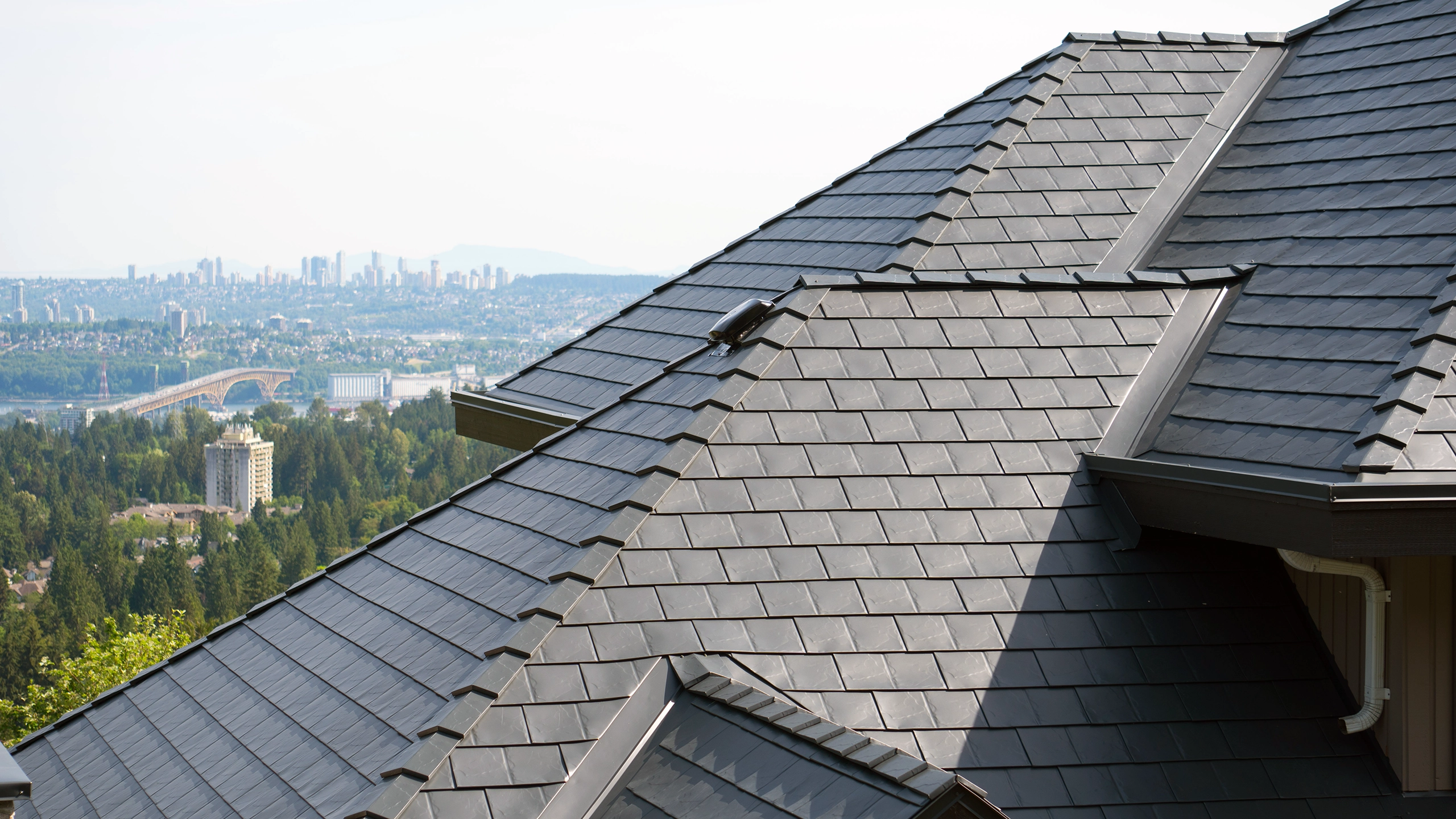 Close-up of a multi-hip deep charcoal Interlock aluminum slate metal roof with complex valleys and ridge caps, overlooking the North Vancouver skyline.