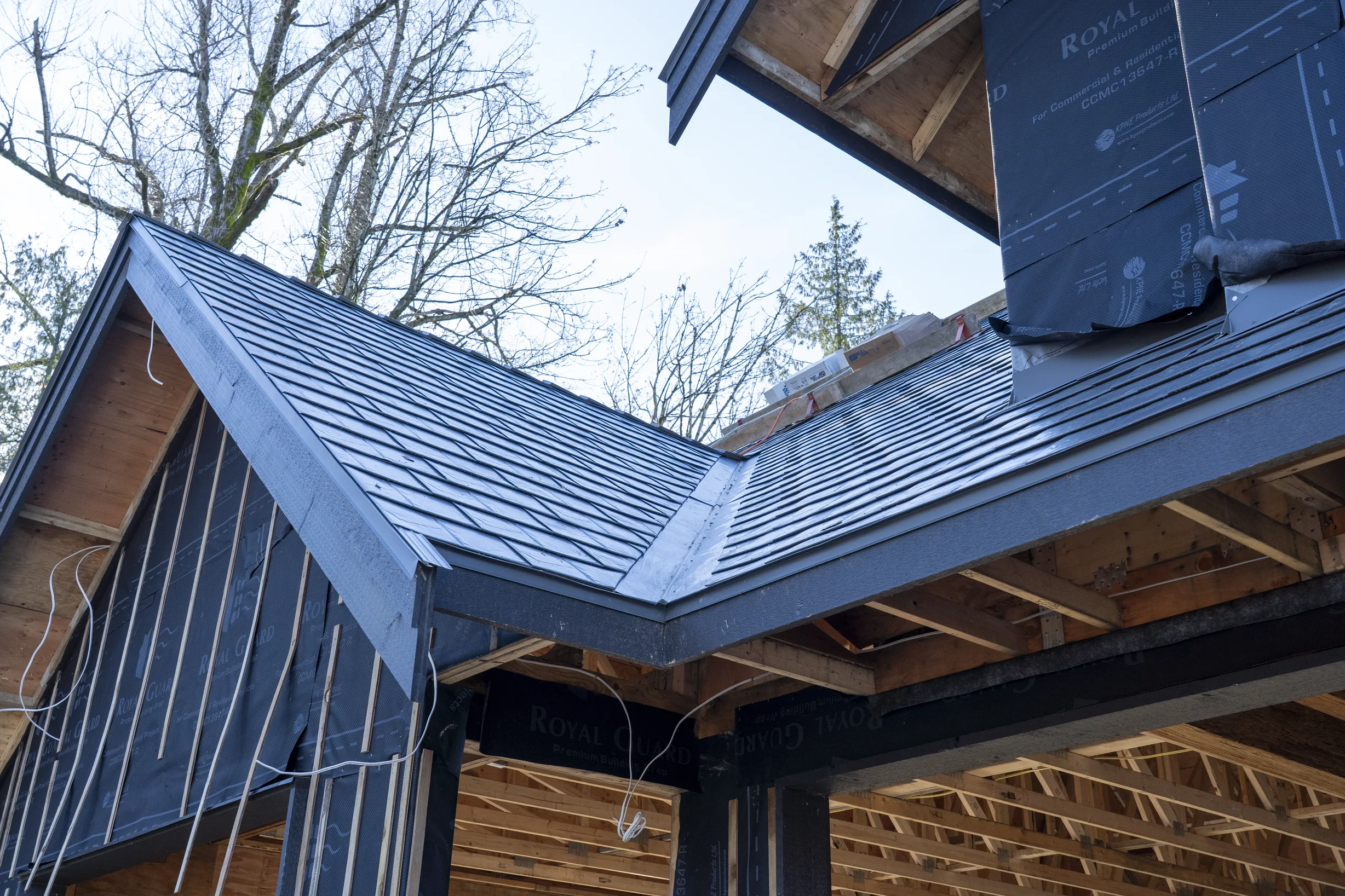 Musket Grey Interlock Slate aluminum roof being installed on a black single-family home with a metal roof in a forested, Abbotsford, British Columbia