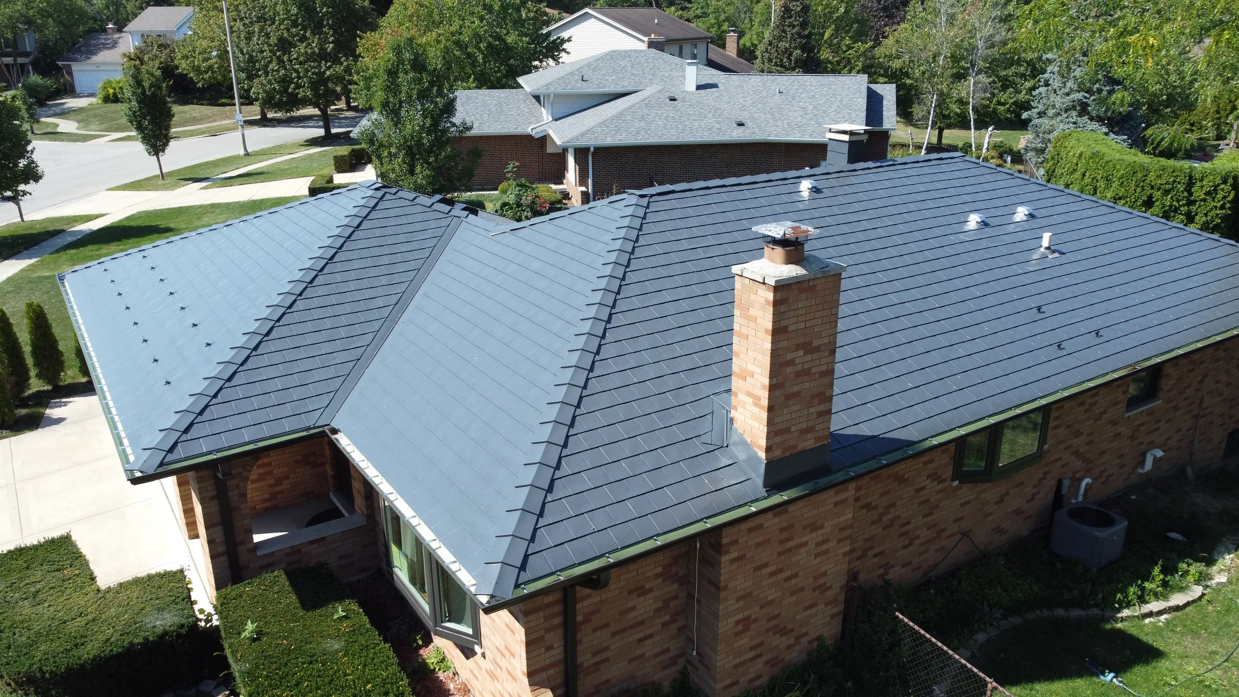 Deep Charcoal Interlock Slate aluminum roof on a red brick home with a metal roof among suburban homes, Westmont, Illinois