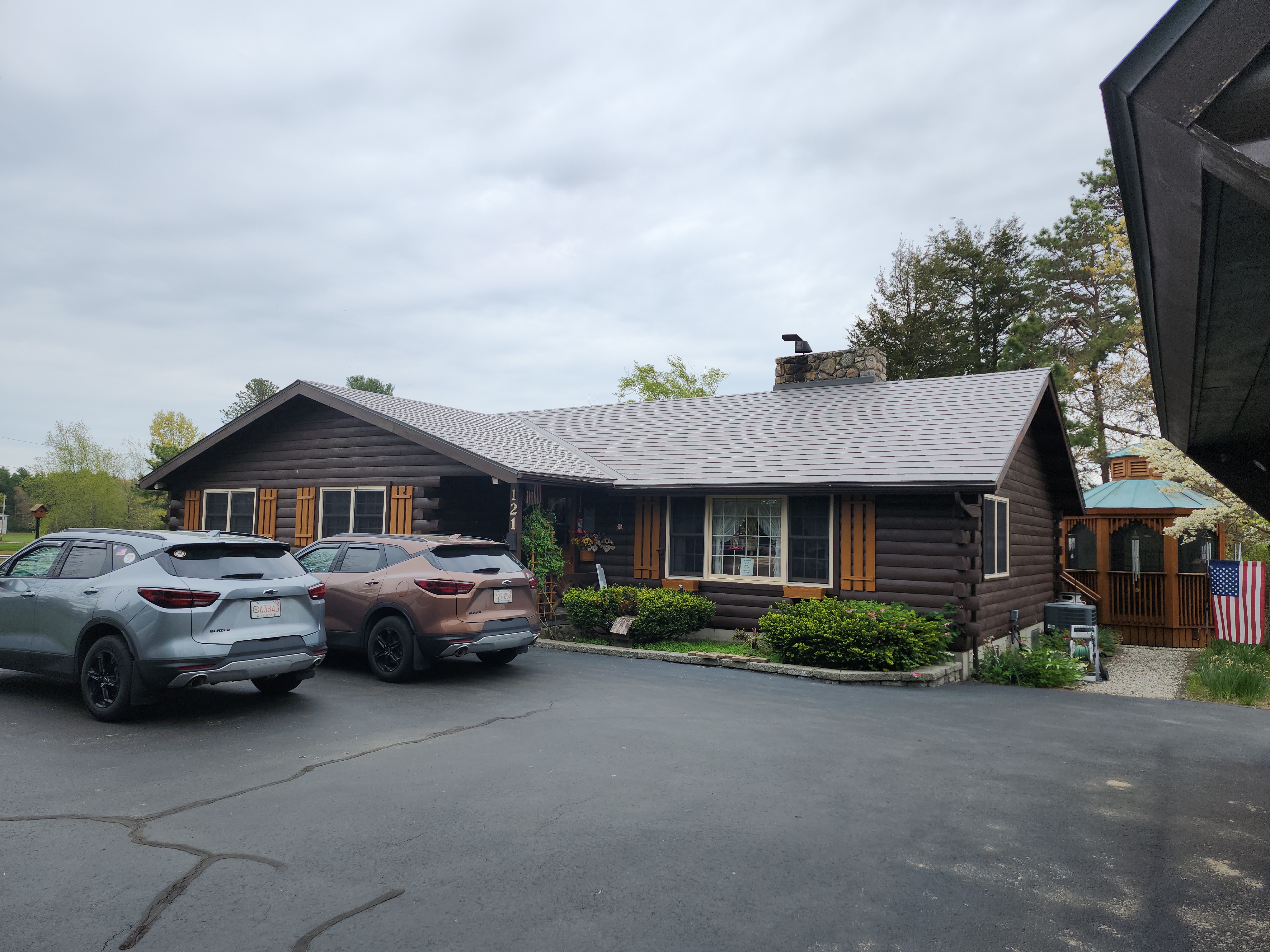 Single-story log-style ranch home in East Freetown MA with Interlock Musket Grey aluminum slate roof, stone chimney, and covered porch with American flag.