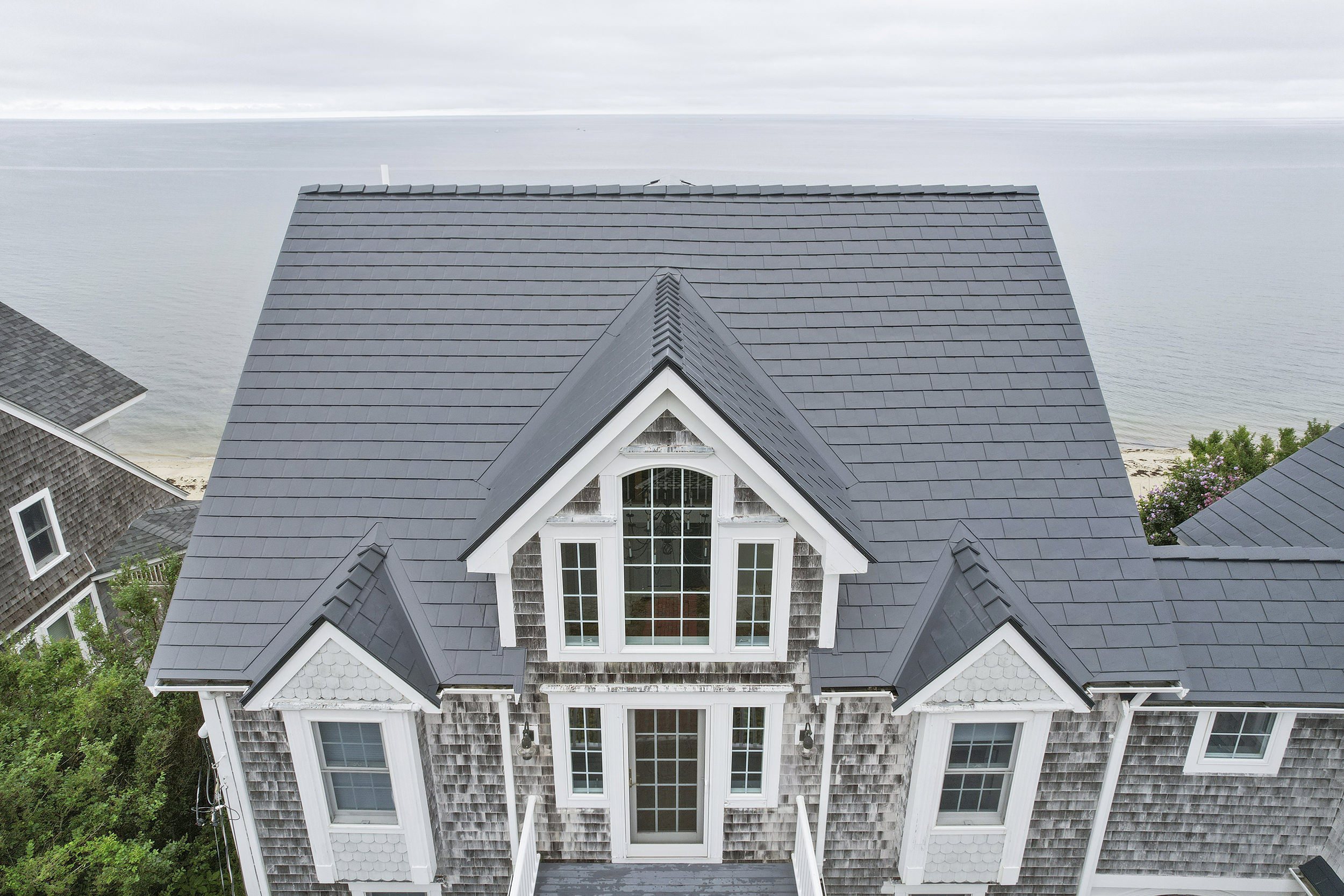 Aerial front view of a Cape Cod beach house with a complex deep charcoal Interlock aluminum slate metal roof featuring multiple dormers and valleys, with ocean backdrop.