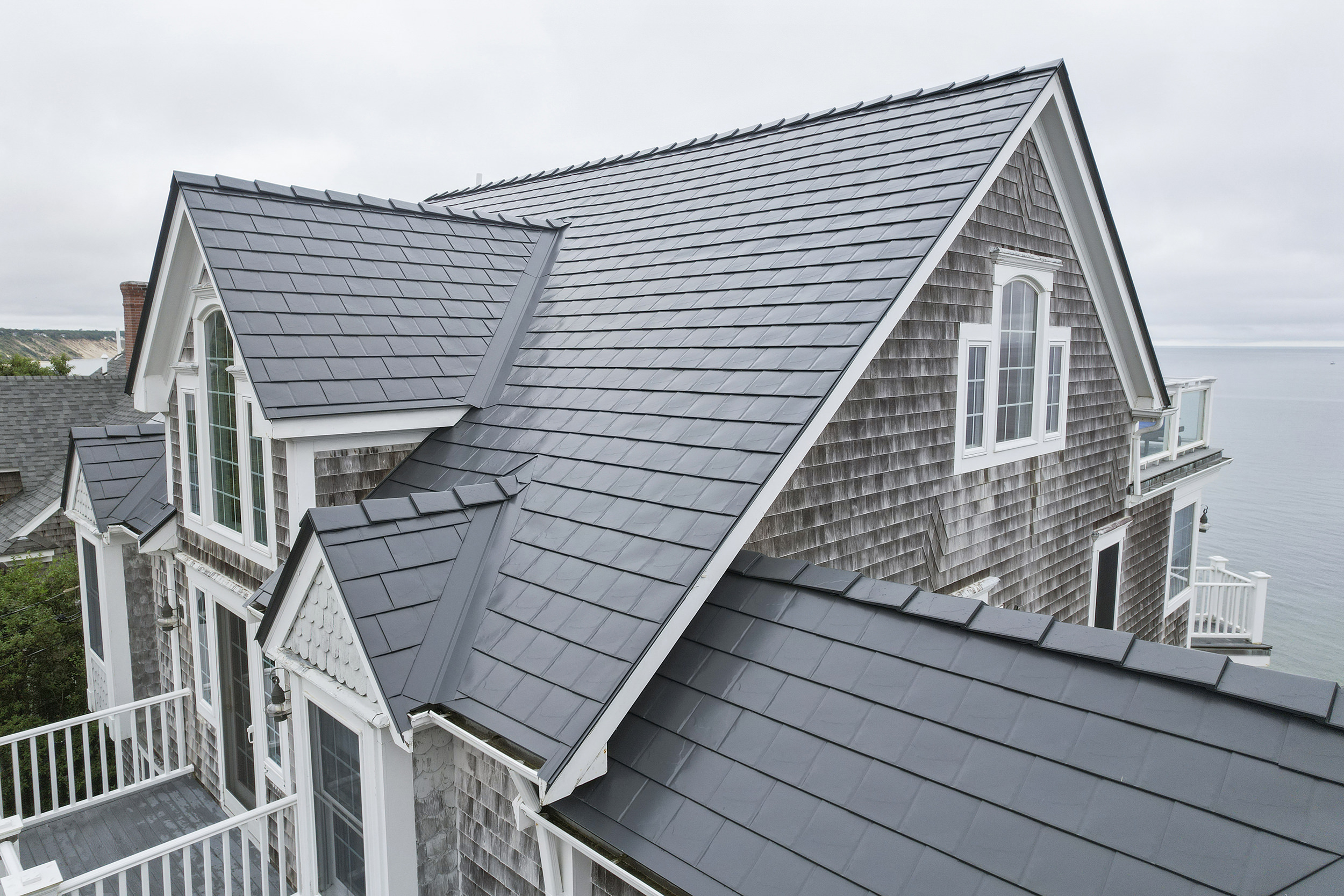 Aerial front view of a coastal shingle-sided home with a complex steep Interlock aluminum slate roof in Deep Charcoal, featuring multiple dormers and valleys.