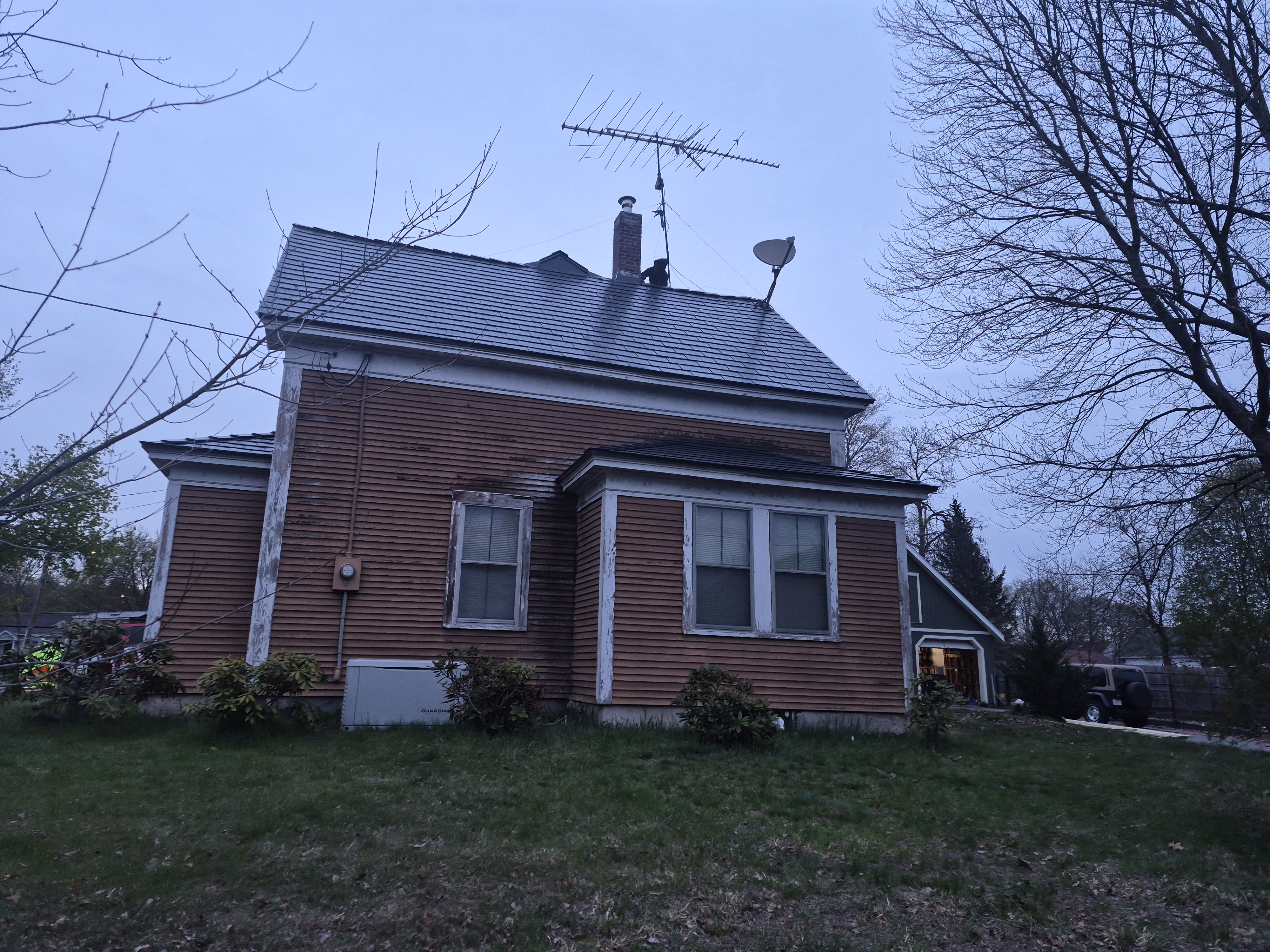 Heritage-style single-family home in Ayer, MA with deep charcoal Interlock aluminum slate mansard roof, brick chimney, and satellite dish at dusk.