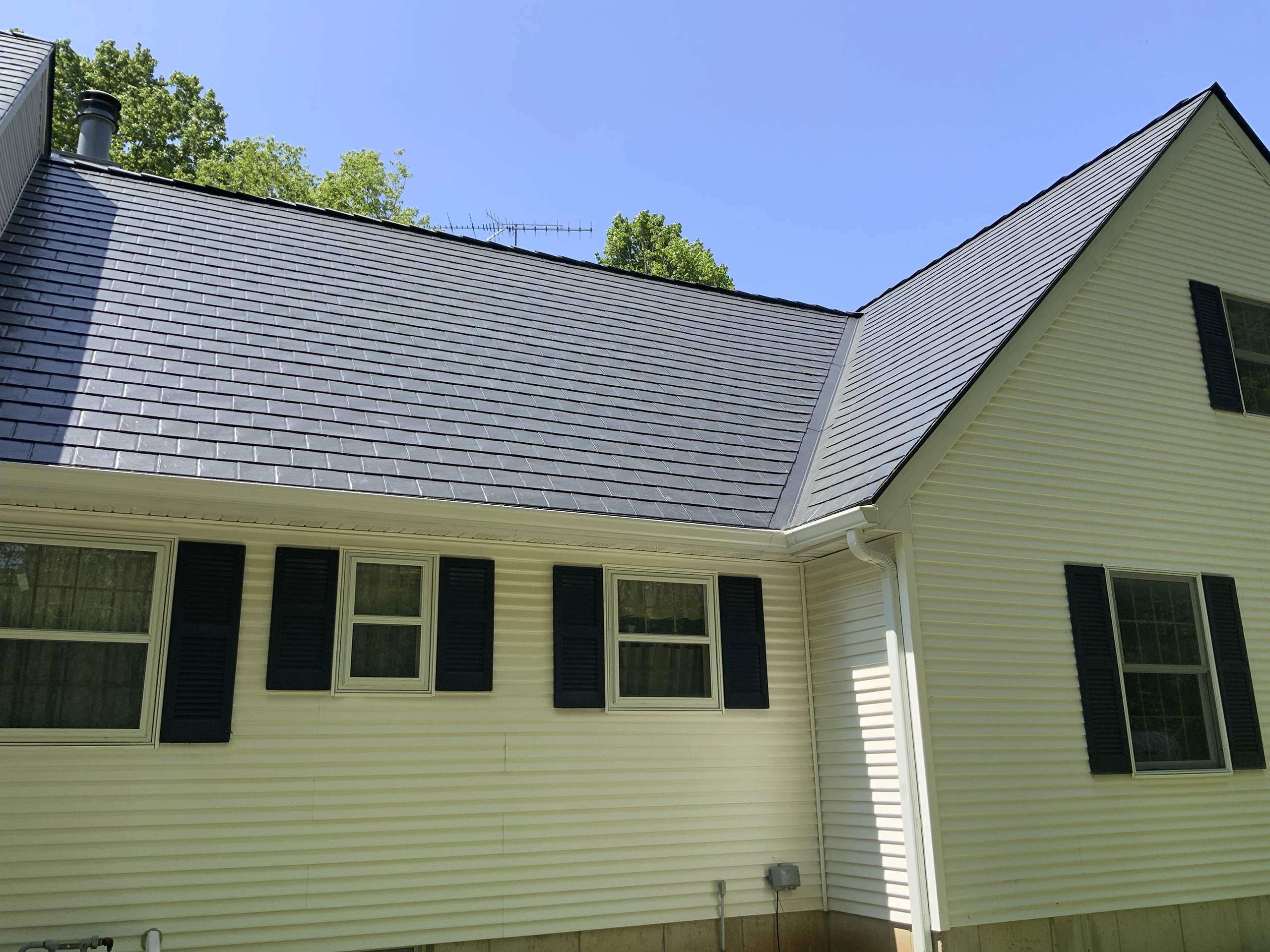 Rear-left view of a Colonial home with Interlock black aluminum slate metal roofing, white vinyl siding, and black shutters under a clear blue sky.