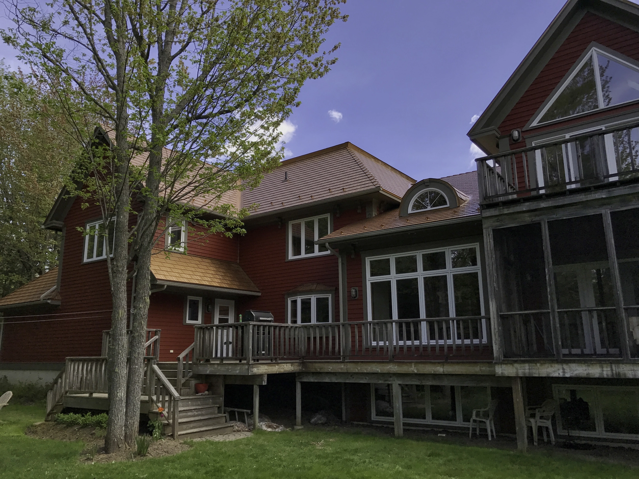 Aged Copper Interlock Cedar Shingle aluminum roof on a red two-storey home with a metal roof among tall pines, Hanwell, New Brunswick