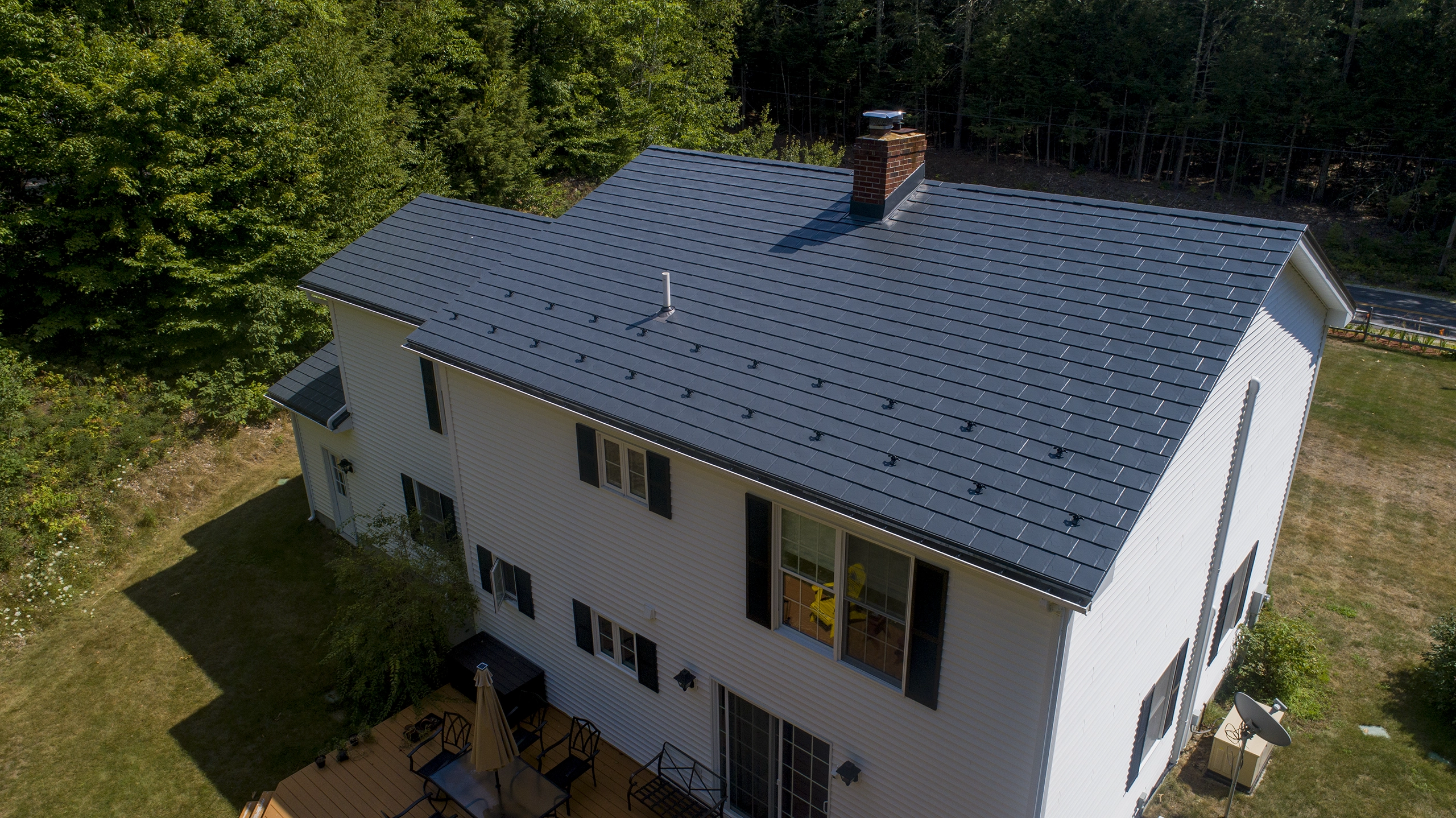 Aerial rear view of a white Colonial home in Webster, NH featuring a deep charcoal Interlock aluminum slate metal roof with snow guards and brick chimney.