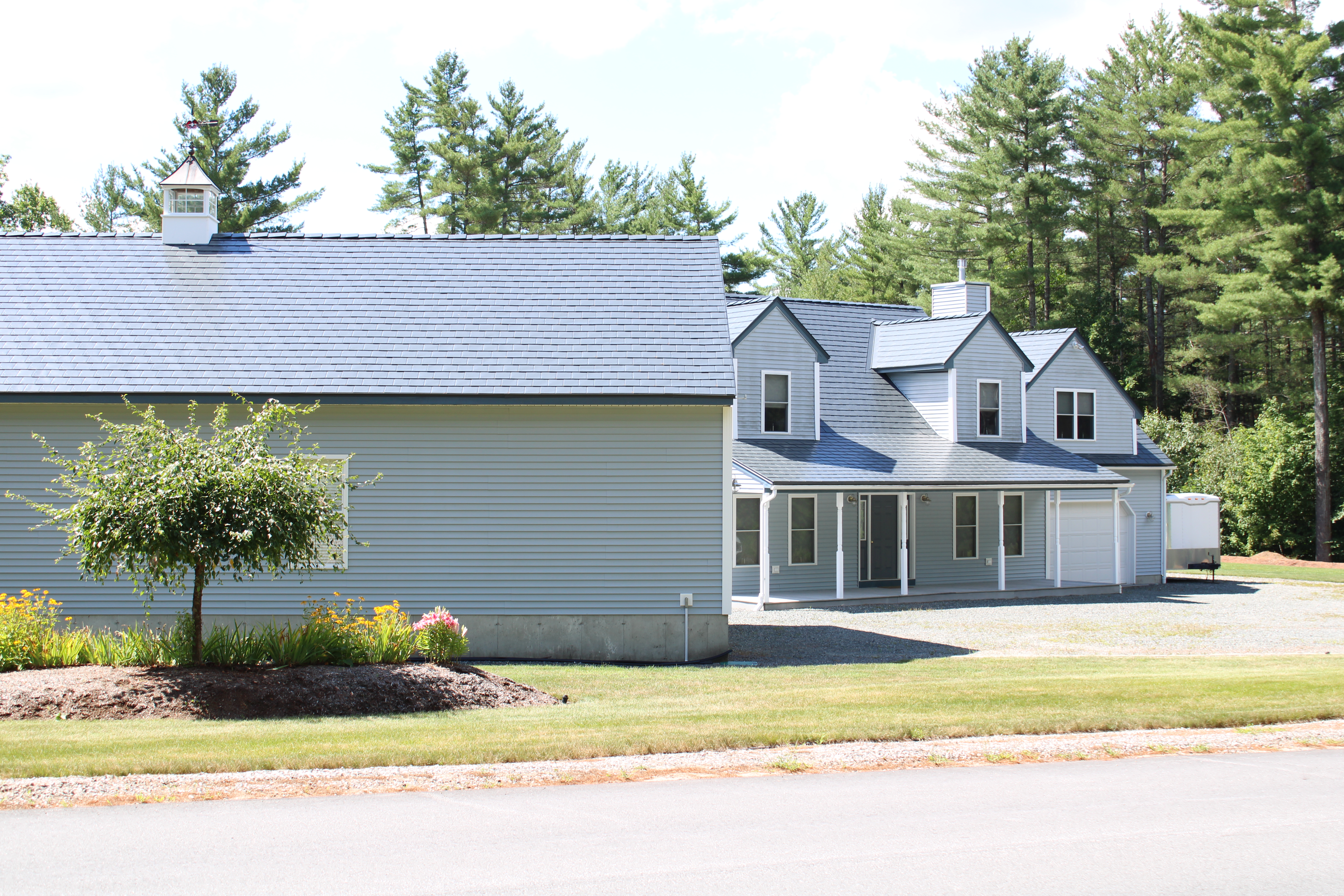 Two-story farmhouse in Thornton NH with Interlock deep charcoal aluminum slate metal roofing, multiple dormers, covered porch, and attached garage amid tall pines.