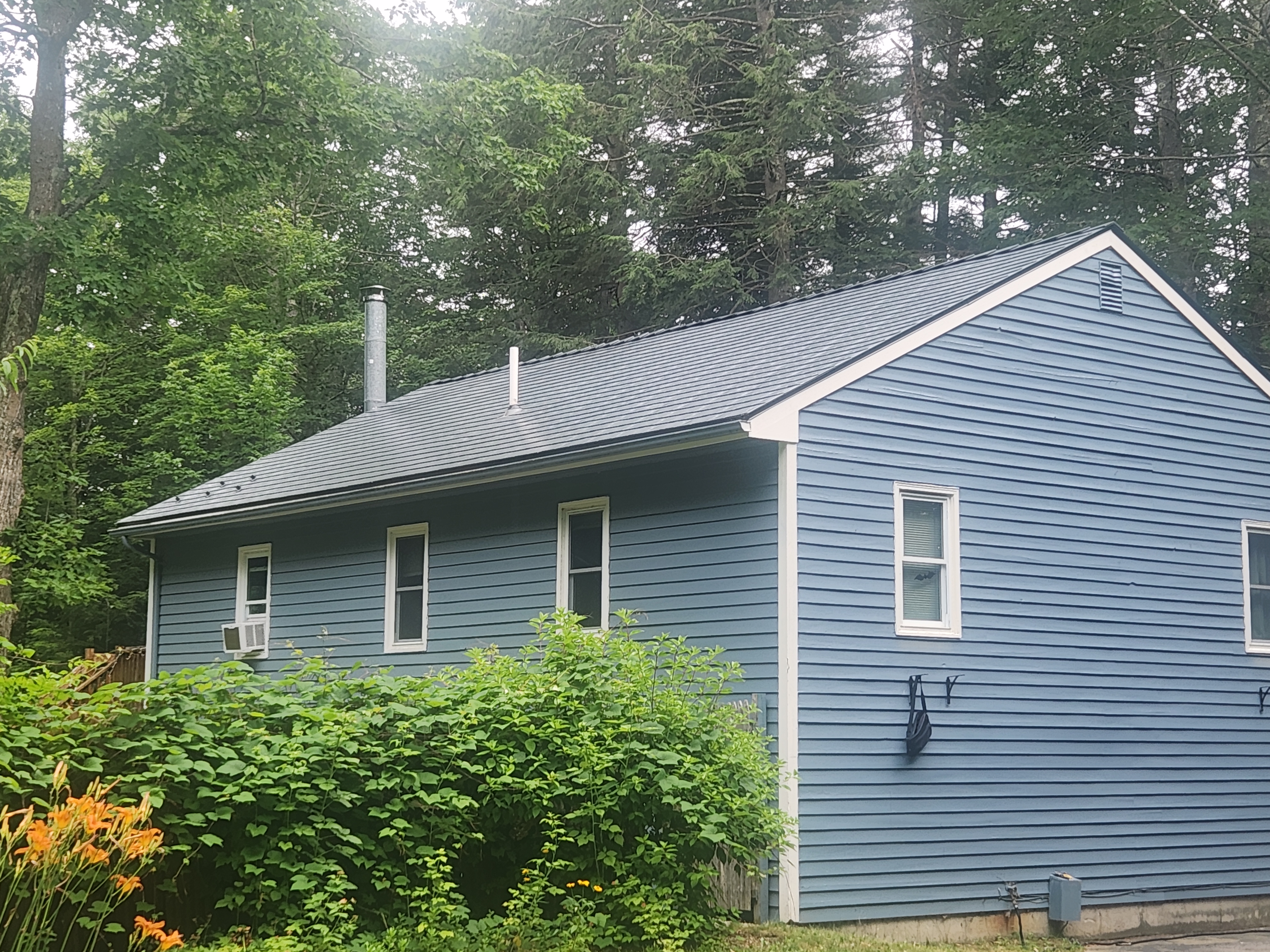 Single-story Ranch home in Bow, NH with a deep charcoal Interlock aluminum slate metal roof, blue vinyl siding, and two vent pipes, set among dense forest.