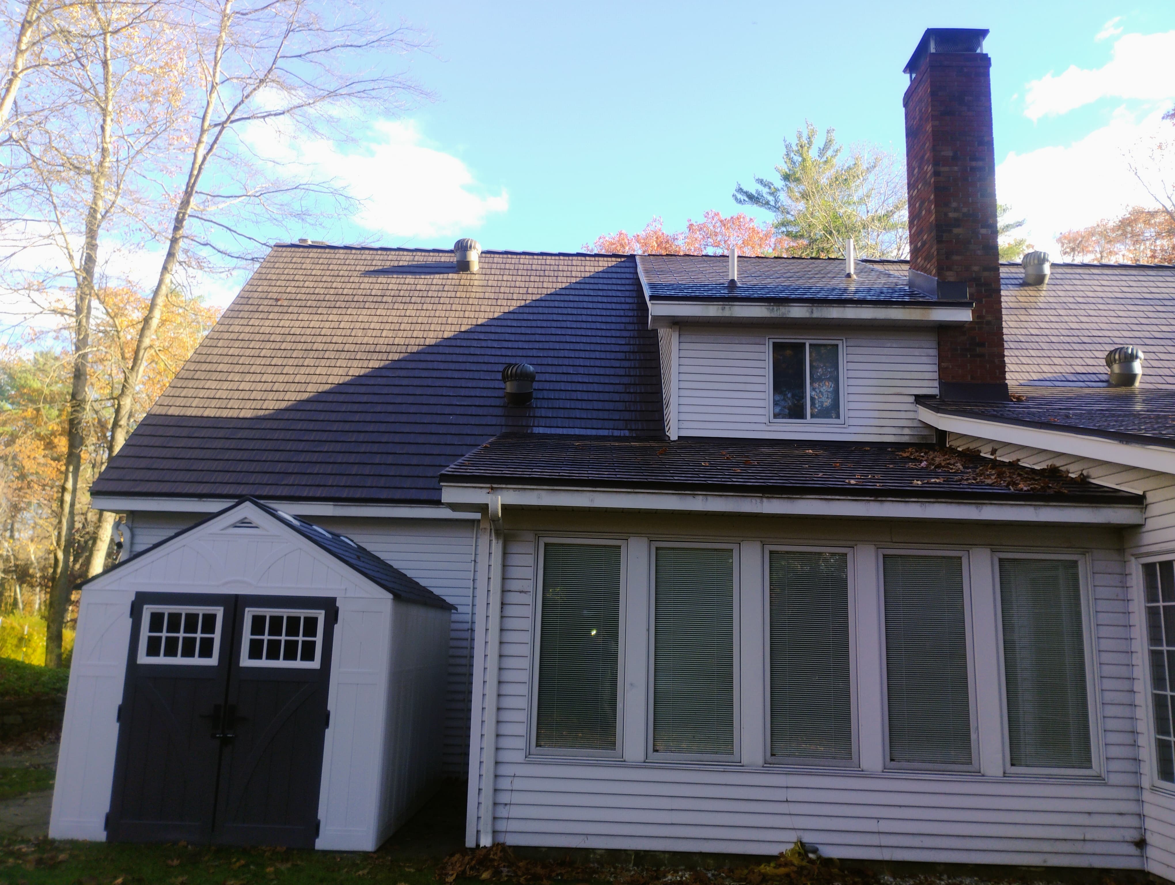 Tudor Brown Interlock aluminum cedar shingle metal roof on a 2-story colonial home with dormer, brick chimney, and multiple roof planes, fall foliage backdrop.