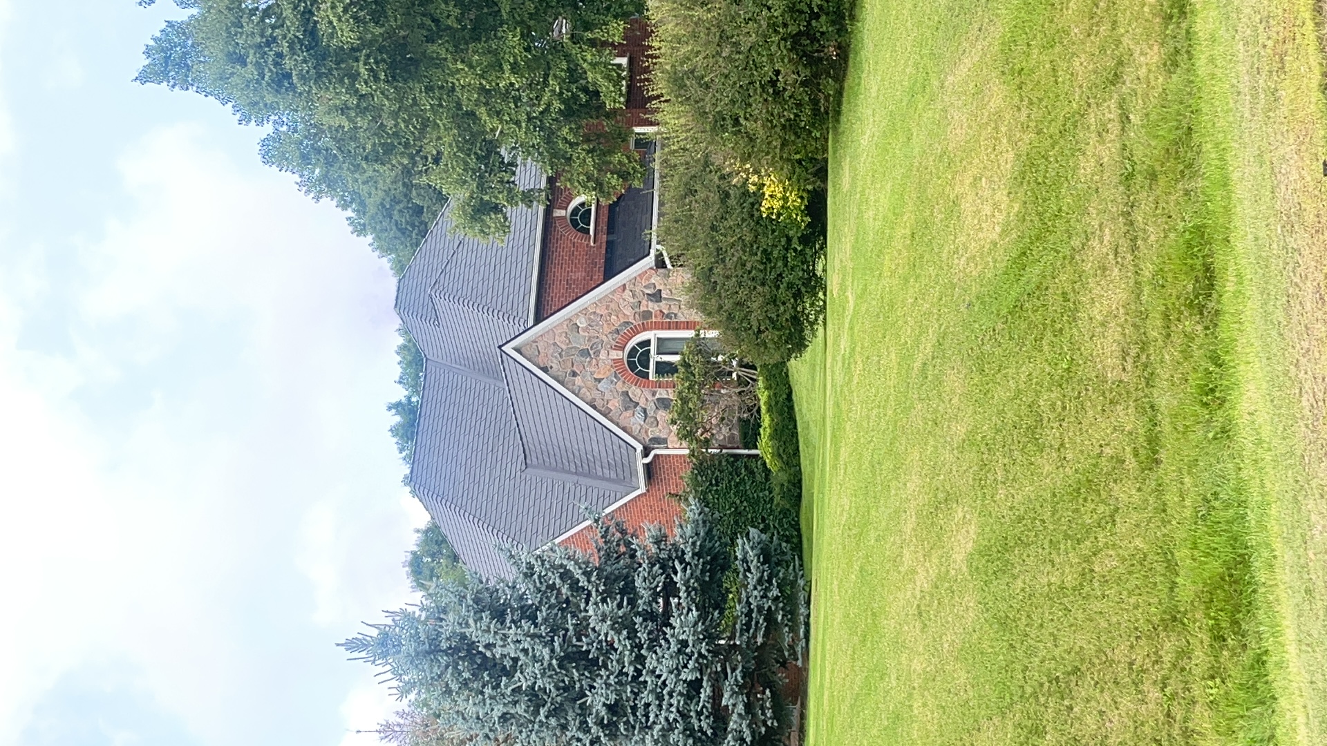 Black Interlock aluminum slate metal roof on a 1.5-storey Tudor-style brick-and-stone home surrounded by mature trees in Campbellville, ON.