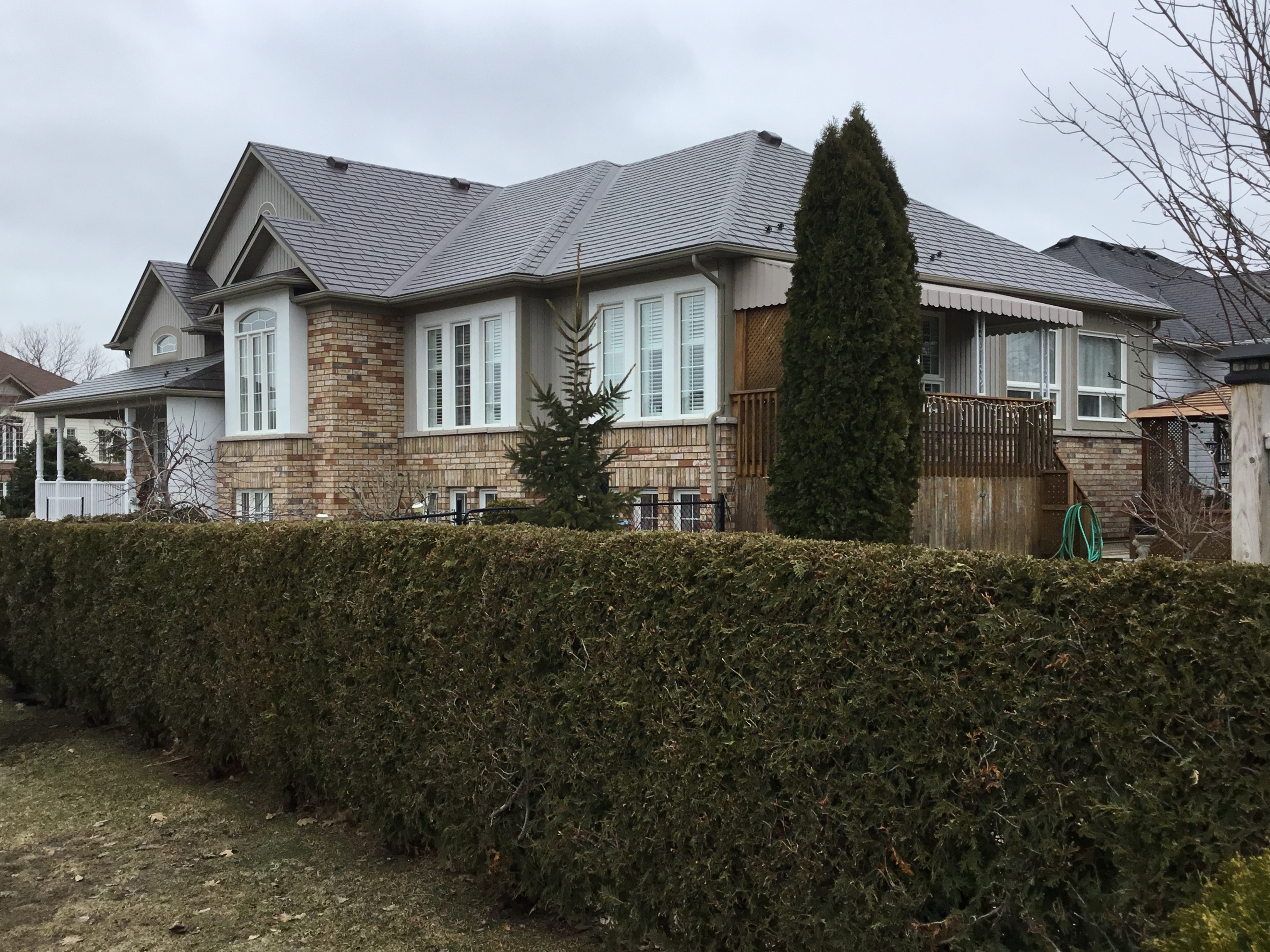 Large single-family home with complex multi-hip Interlock aluminum slate roof in Tudor Brown, brick exterior, viewed from front-right across a trimmed cedar hedge.