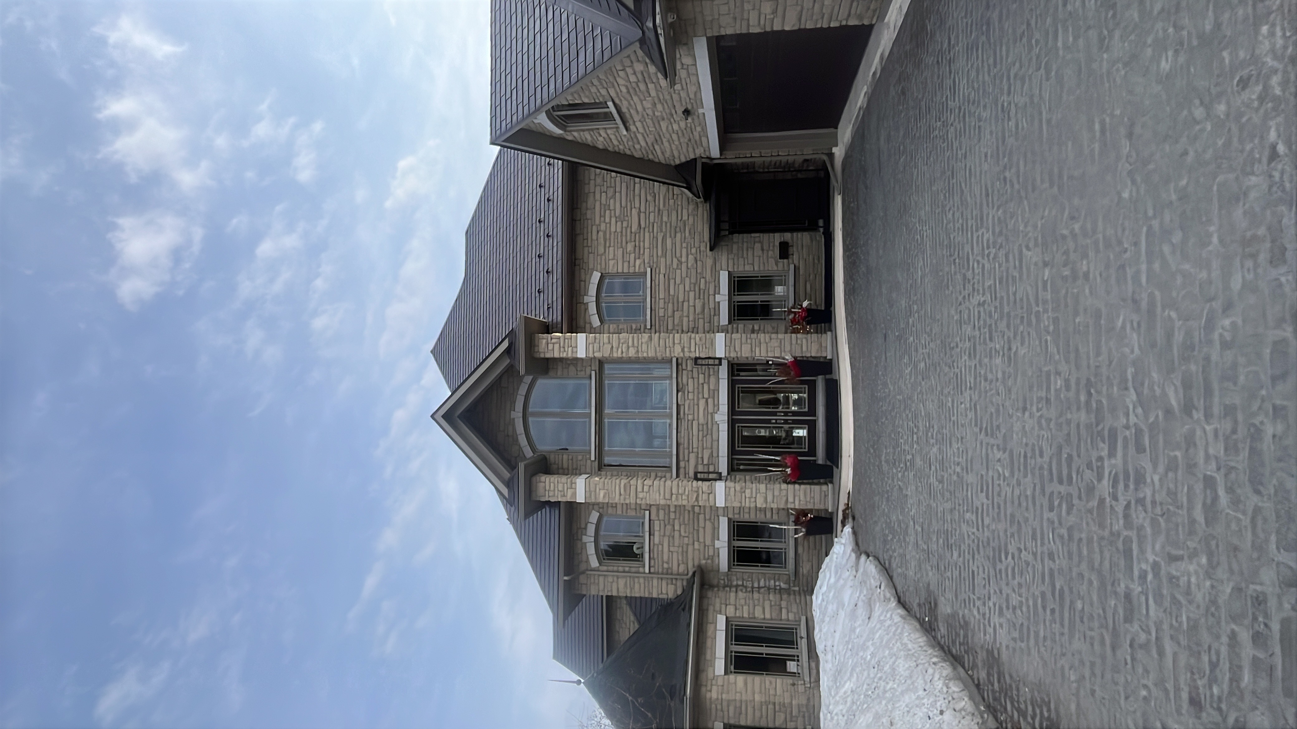 Two-storey Tudor-style stone home in Sudbury ON with complex multi-hip Interlock Tudor Brown aluminum slate roof and cobblestone driveway in winter.