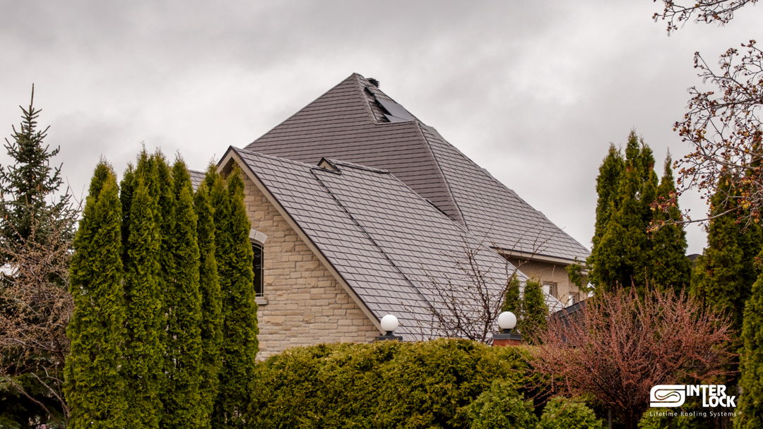 Large Tudor-style stone home in Sudbury ON featuring a steeply pitched multi-hip Interlock aluminum slate roof in Tudor Brown, surrounded by mature evergreens.