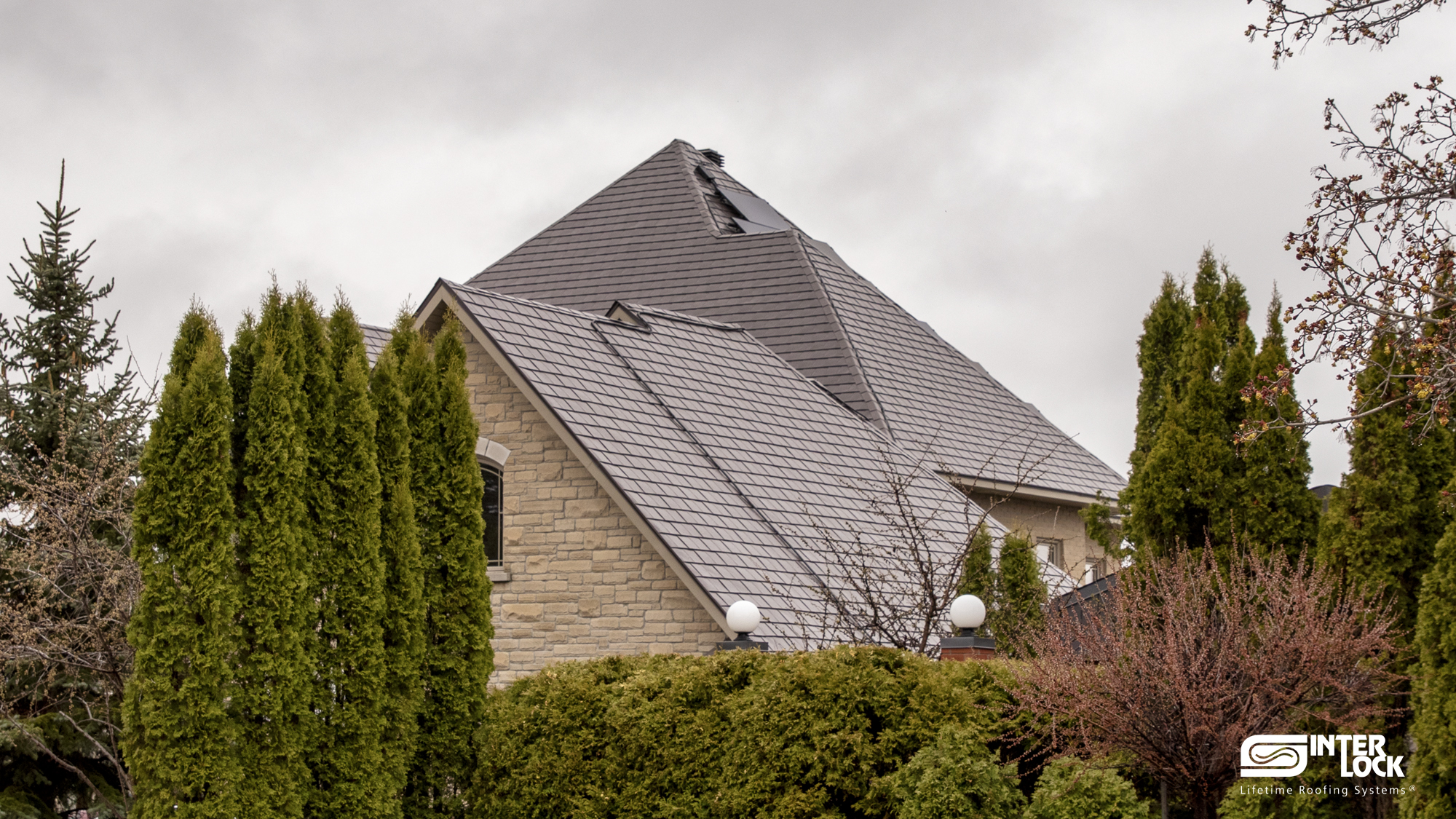 Large two-storey stone home with a steeply pitched multi-hip Interlock aluminum slate roof in Tudor Brown, featuring skylights and complex valleys.