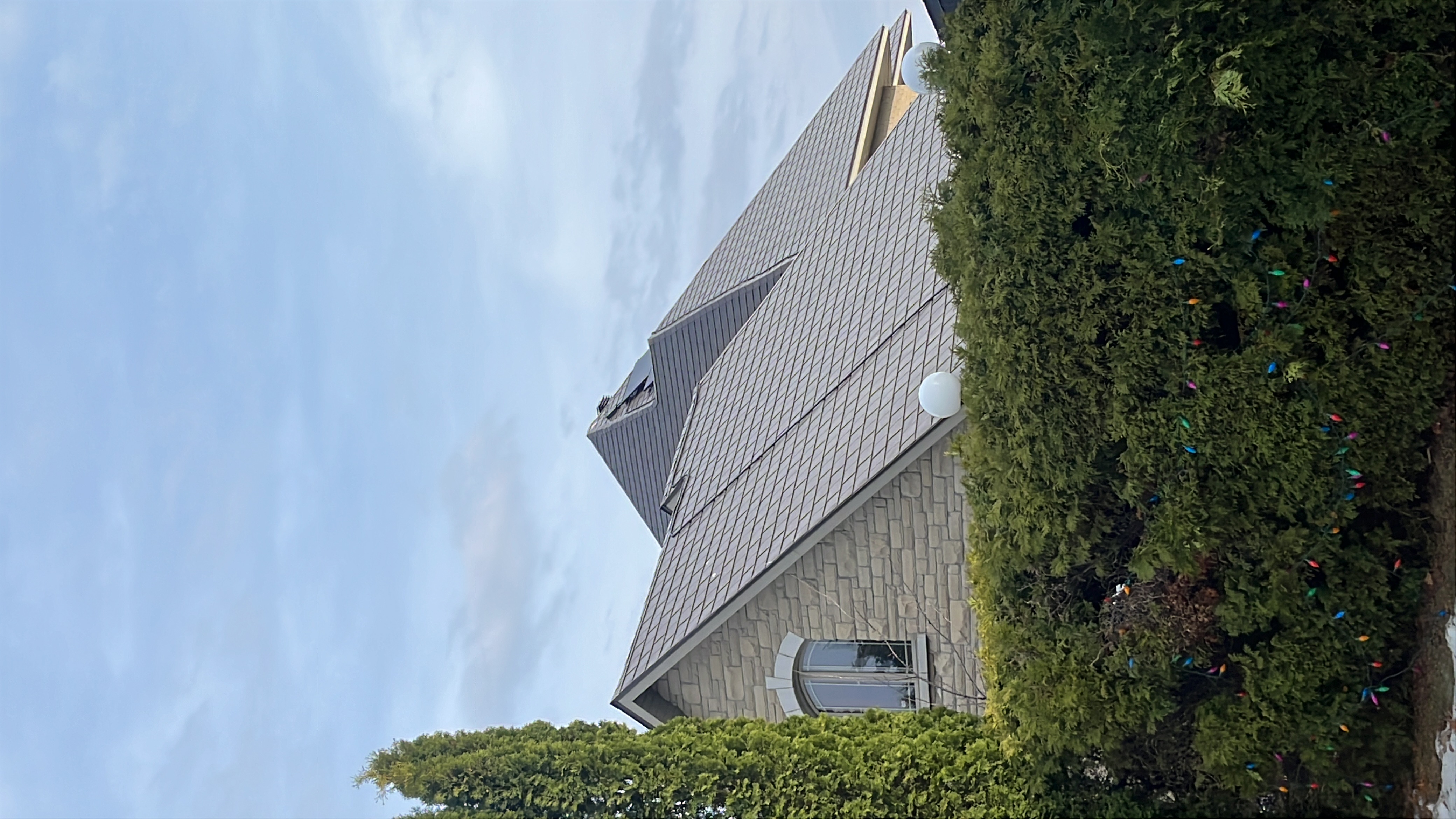 Steep multi-hip Interlock aluminum slate roof in Tudor Brown on a stone two-storey home in Sudbury, ON, framed by tall cedars and holiday lights.