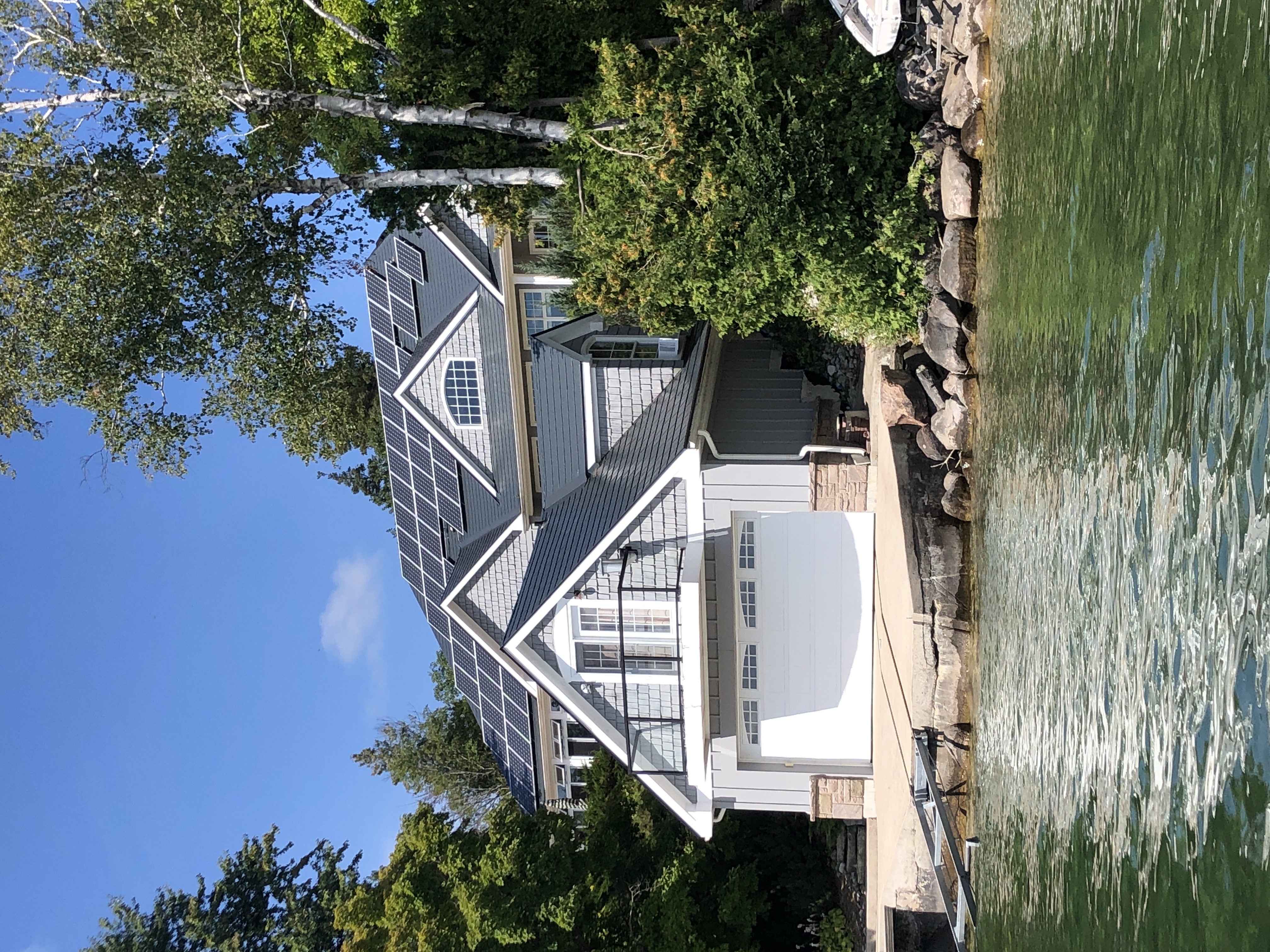 Craftsman lakefront home with deep charcoal Interlock aluminum slate roof, multiple solar panels, complex multi-gable profile, and boathouse garage on rocky shoreline.