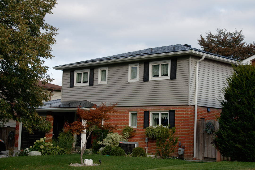 Two-storey suburban home with black Interlock aluminum slate metal roofing, red brick lower facade, grey vinyl siding, and multi-hip roof profile in Scarborough, ON.