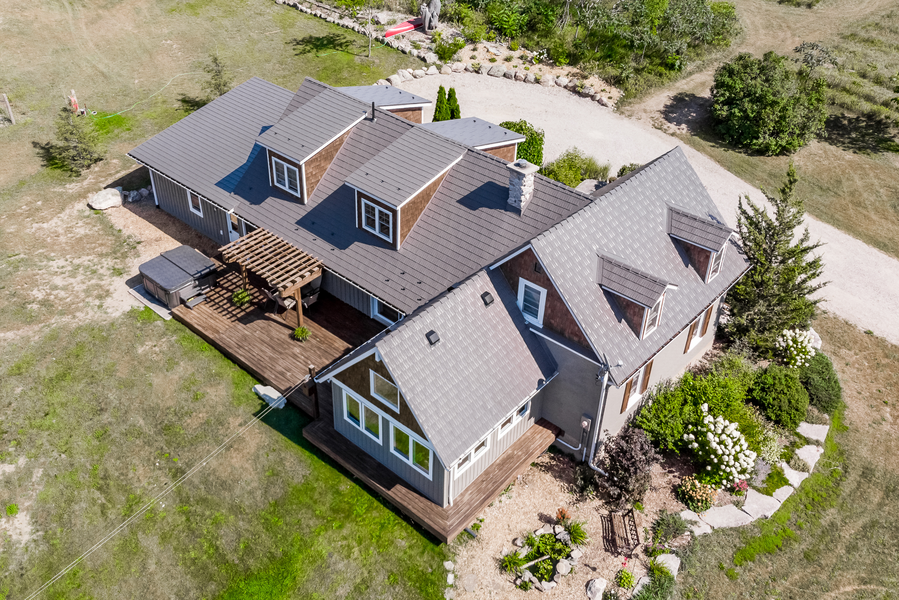 Aerial rear view of a 1.5-storey rural home in Midland, ON with Interlock weathered cedar shingle metal roofing, multiple dormers, chimney, and wood deck.