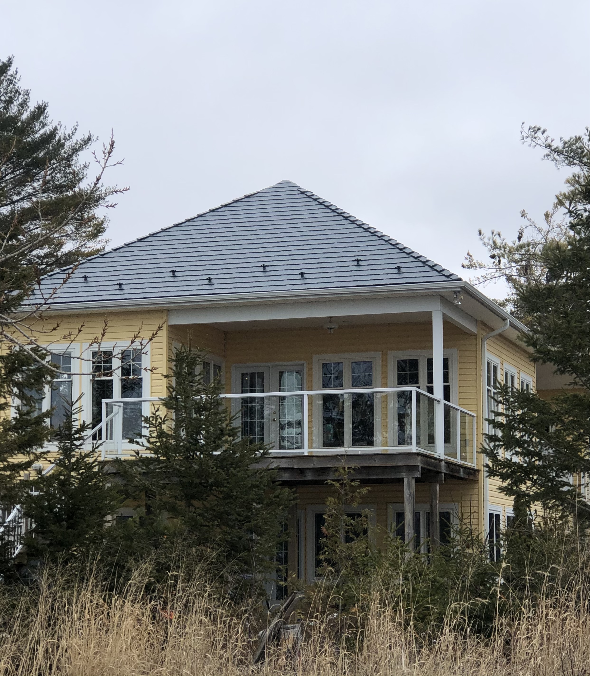 Two-storey yellow cottage with a hip-profile deep charcoal Interlock cedar shingle metal roof, snow guards, and a covered upper deck balcony, set among coastal grasses and pines.